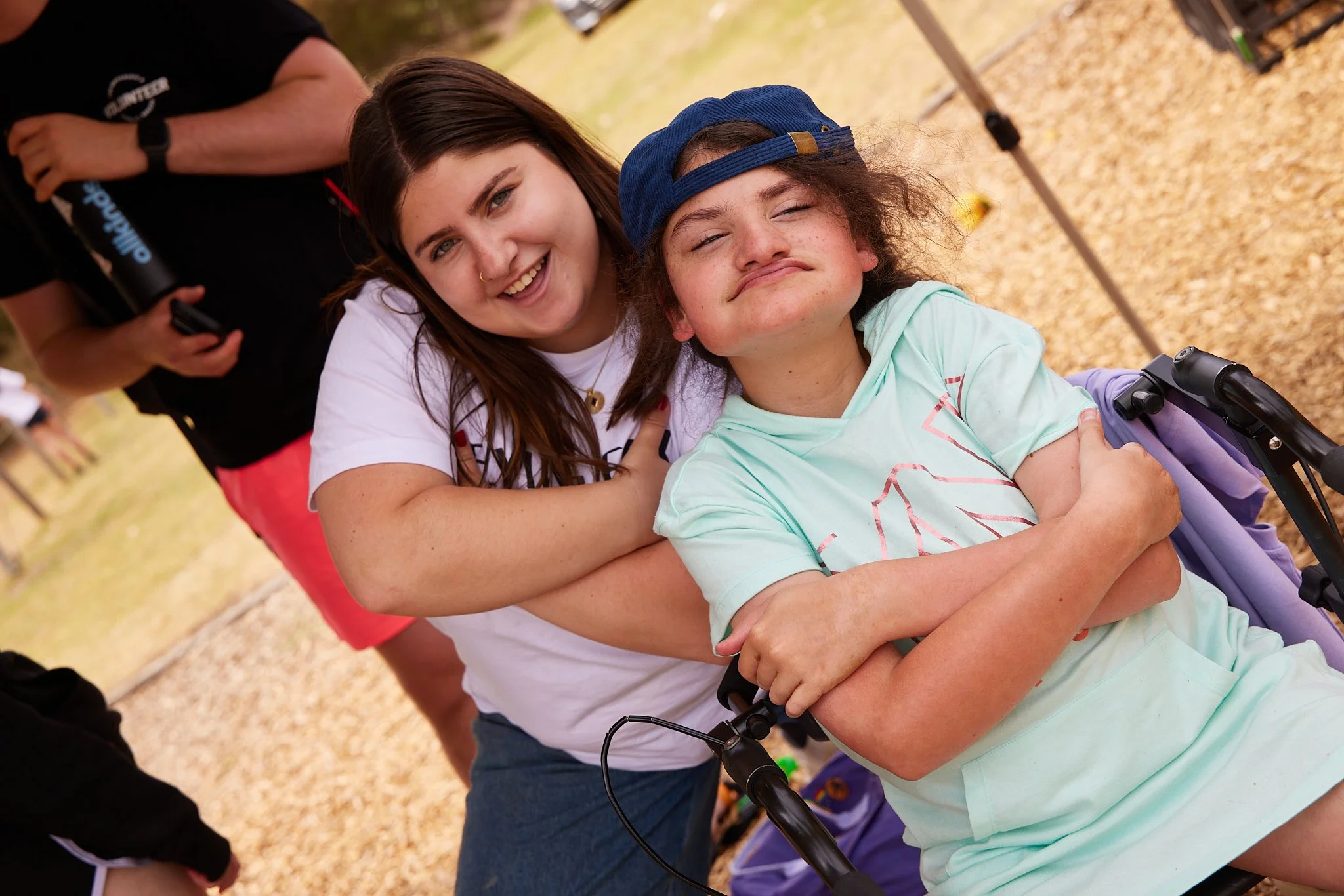 Two young women smiling and hugging outdoors, one pushing a wheelchair with a girl in it wearing a blue cap.