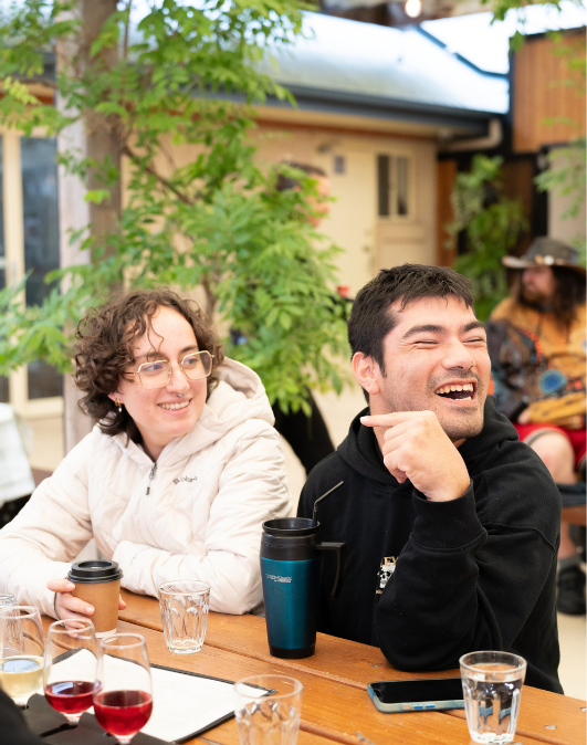 Two friends sitting at a wooden table outdoors, smiling and laughing. The woman on the left has curly hair, glasses, and is wearing a light-colored jacket. The man on the right has dark hair, a beard, and is wearing a black hoodie. There are glasses of wine, cups of coffee, and a smartphone on the table. In the background, there are plants, a house, and a person in a hat.