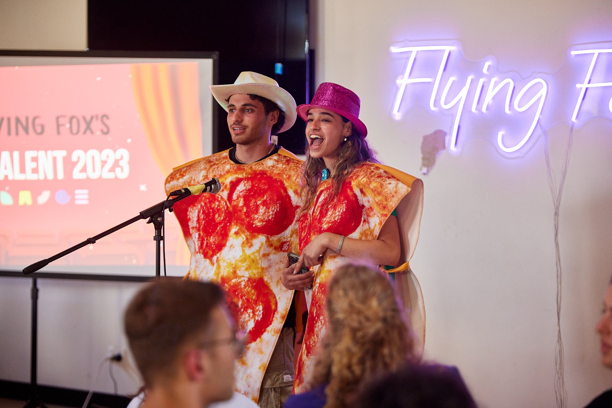Two people dressed in pizza slices costume standing on stage at a talent show, with a man wearing a cowboy hat and a woman wearing a pink glitter ha. Neon sign reads 'Flying Fox' and a screen in the background says 'Flying Fox's Talent 2023'.