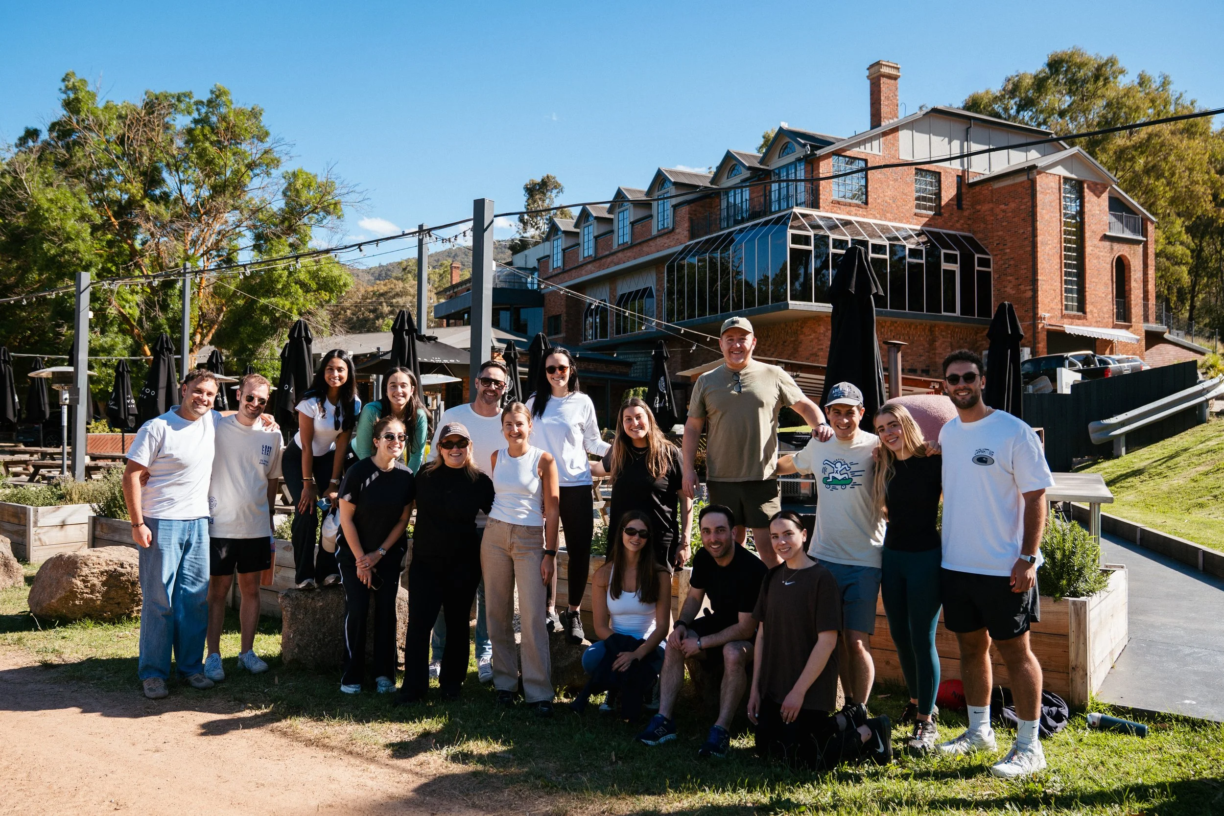 Group of people smiling outdoors in front of a large brick building with glass windows and black umbrellas, on a bright sunny day.