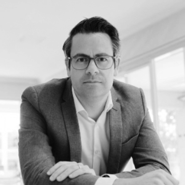 A man with glasses and dark hair wearing a suit jacket and white shirt, looking serious, sitting at a desk in a bright, modern office.