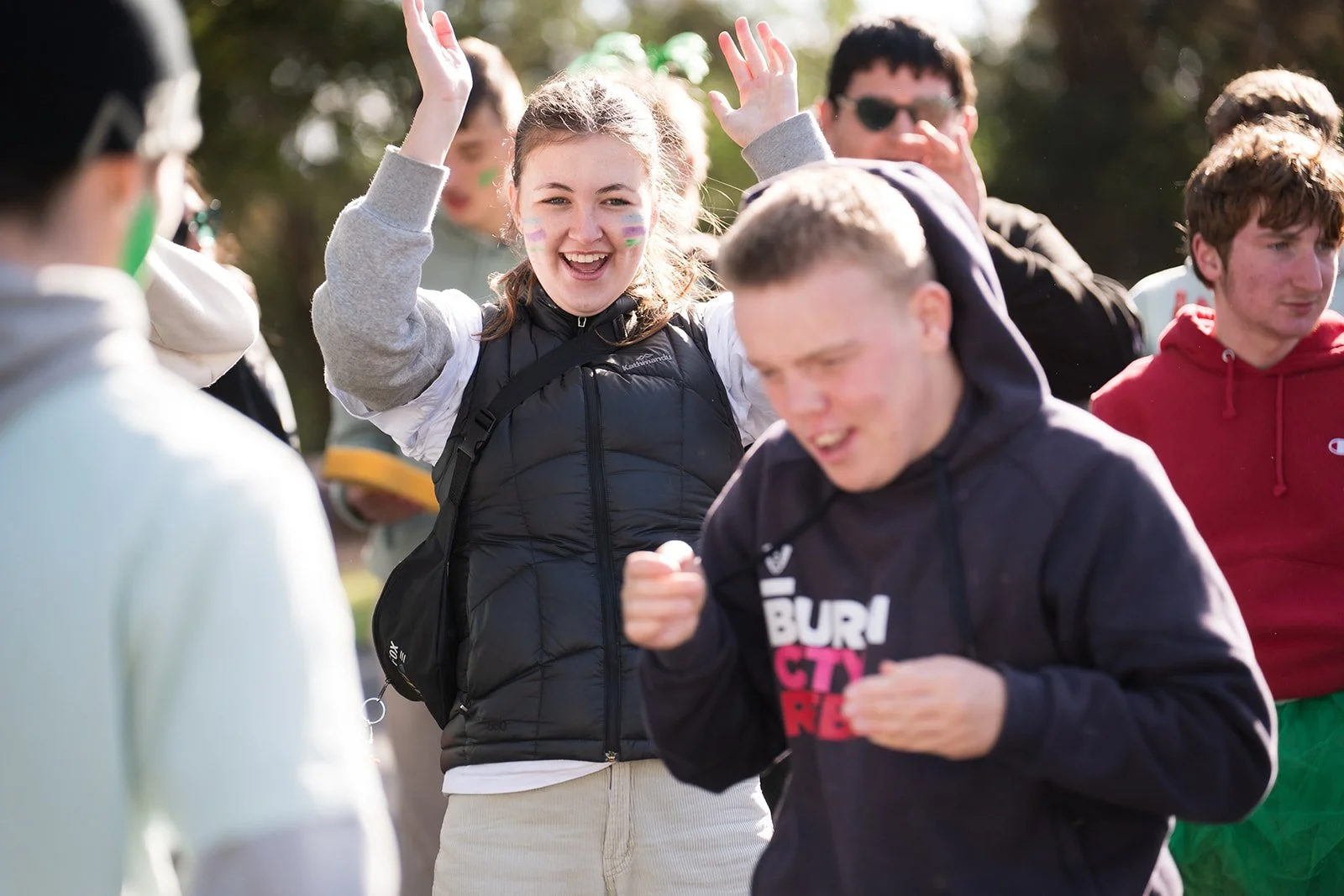 A group of young people outdoors, some smiling and raising their hands, dressed in casual and athletic clothing, enjoying a sunny day, likely during a social or recreational event.