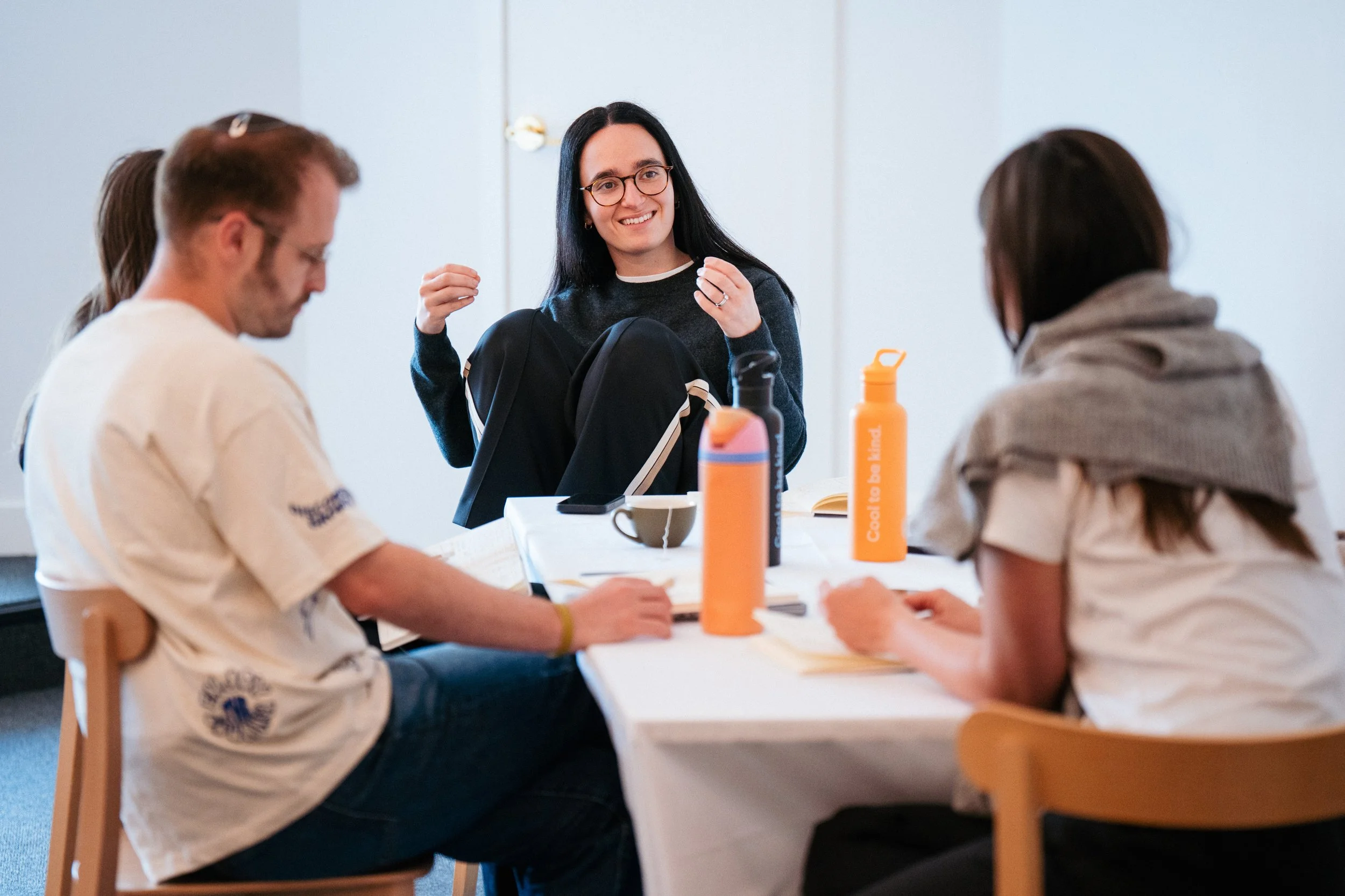 A group of four young people sitting at a meeting table, one woman smiling and talking, with water bottles and notebooks on the table.