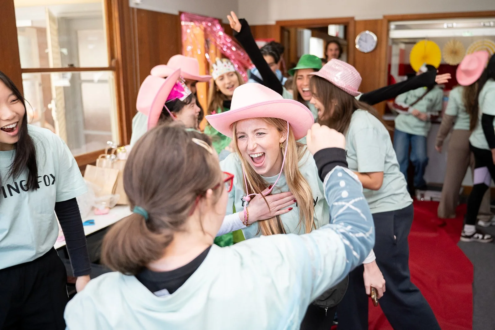 A group of women celebrating, wearing colorful hats and joyful expressions, with some women hugging and others cheering in a festive indoor setting.