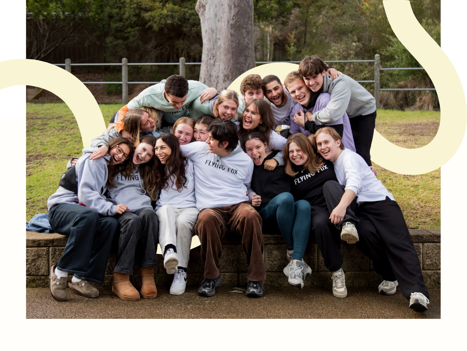 A group of young people sitting on a brick ledge outdoors, laughing and hugging, with trees and a fence in the background.