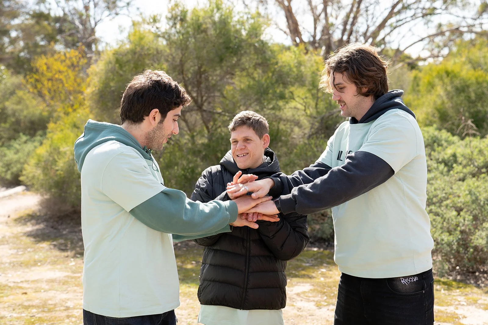 Three young men with dark hair outdoors on a sunny day, engaged in a friendly handshake or gesture of camaraderie, with trees and greenery in the background.