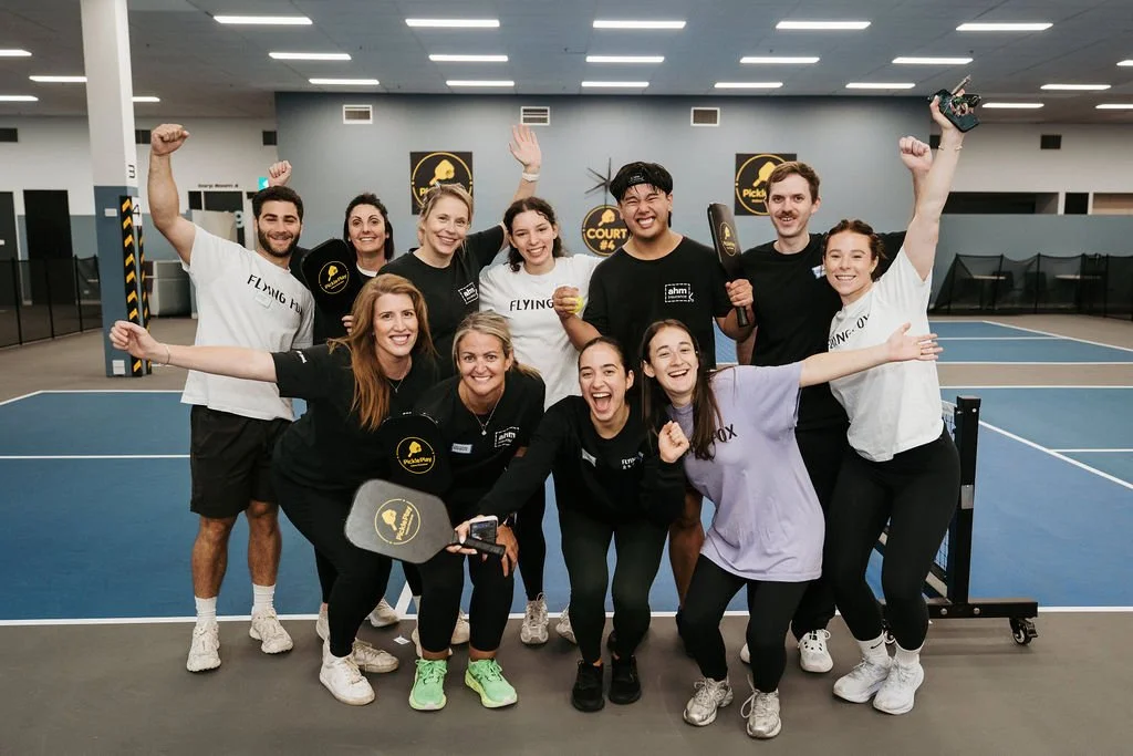 Group of people celebrating on an indoor tennis court, some holding paddles and a trophy, smiling and making celebratory gestures.
