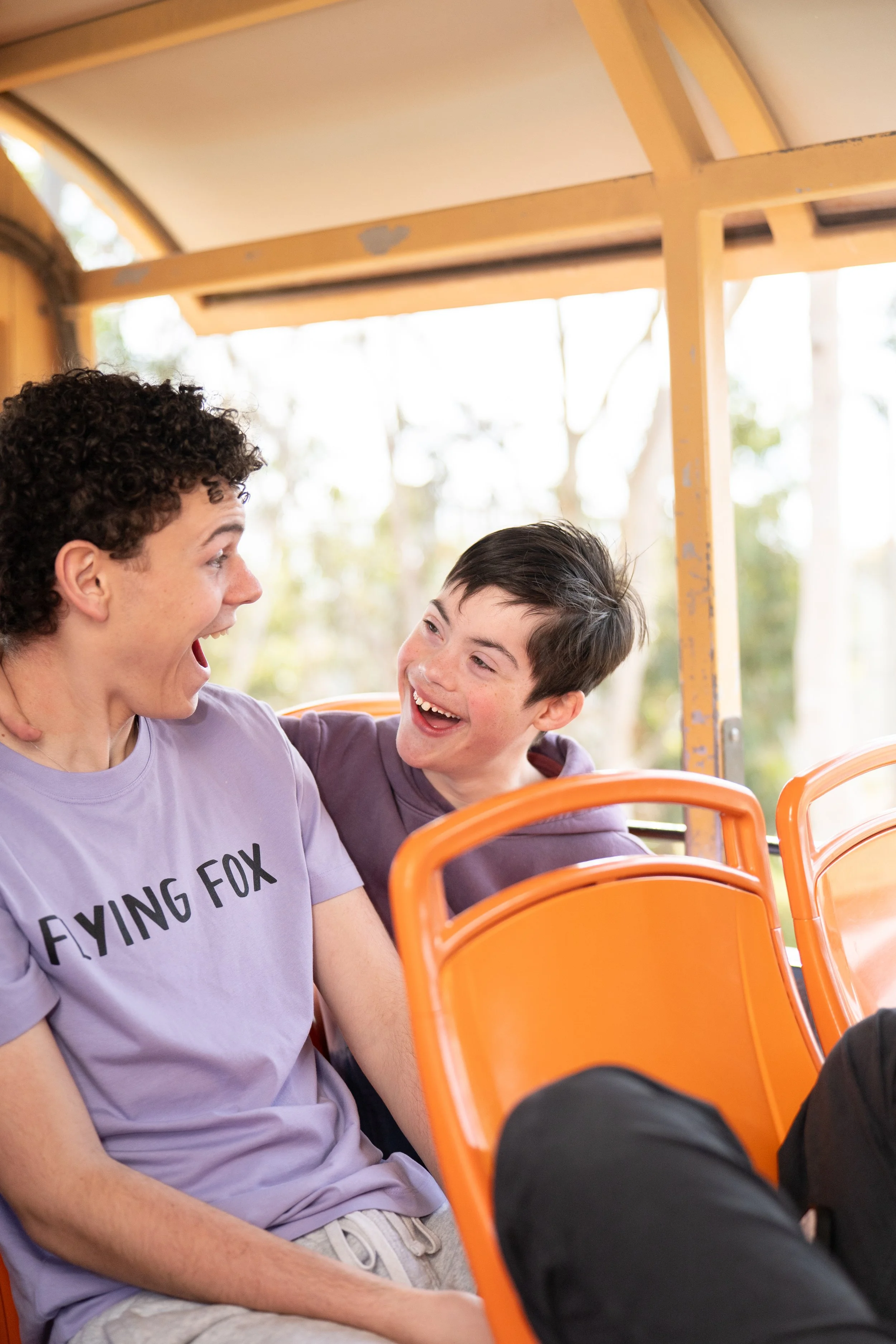 A young man and a boy are sitting on a bright orange bus, both smiling and looking at each other with joy. The  is wearing a lavender T-shirt with the words Flying Fox, and the boy is wearing a purple hoodie. They are inside the bus with a natural background visible through the windows.