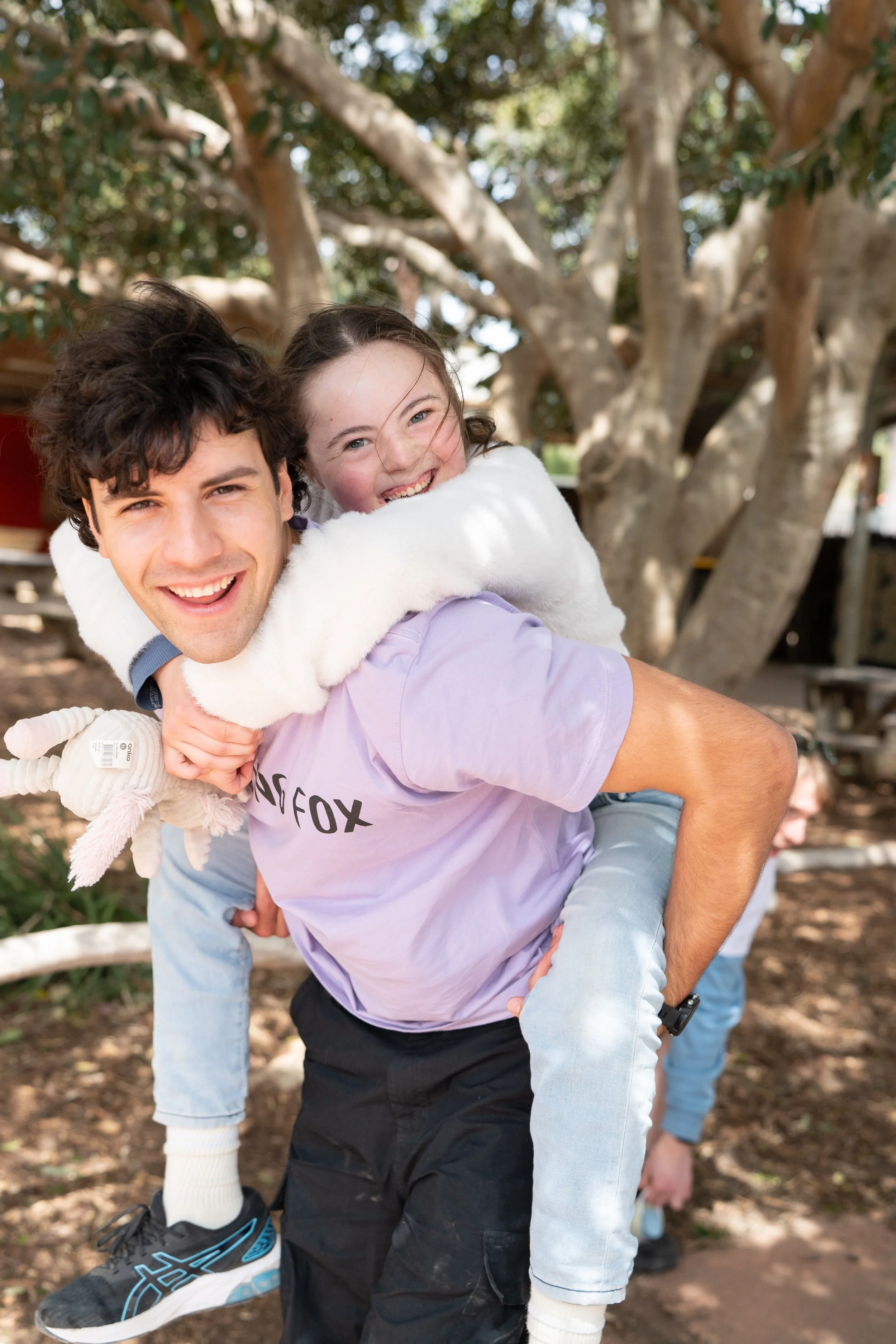 A young man giving a piggyback ride to a smiling girl outdoors near a tree, with another person partially visible in the background.