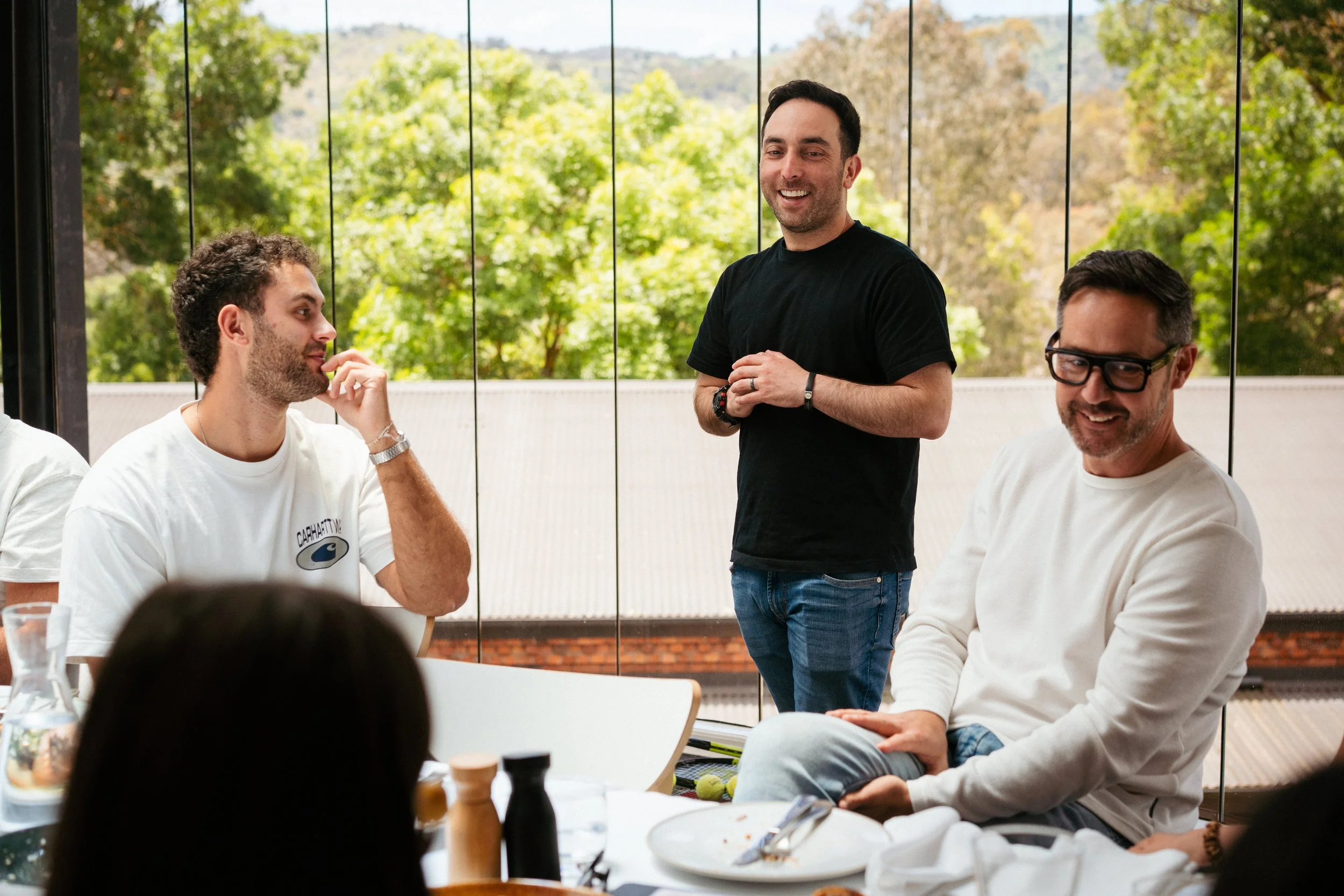 Group of men at a table having a conversation, with one standing and smiling, outside view with green trees in the background.