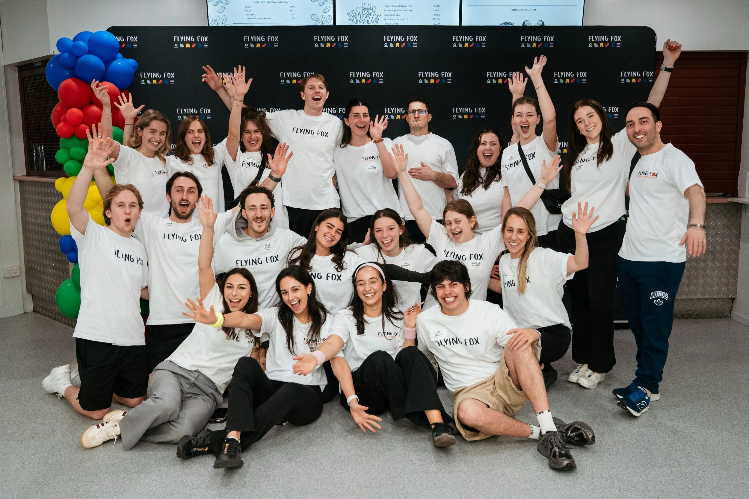 A group of young adults posing for a photo in front of a black backdrop with the Flying Fox logo, with colorful balloons on the left side. They are all smiling, some waving, and wearing white T-shirts with the Flying Fox logo.