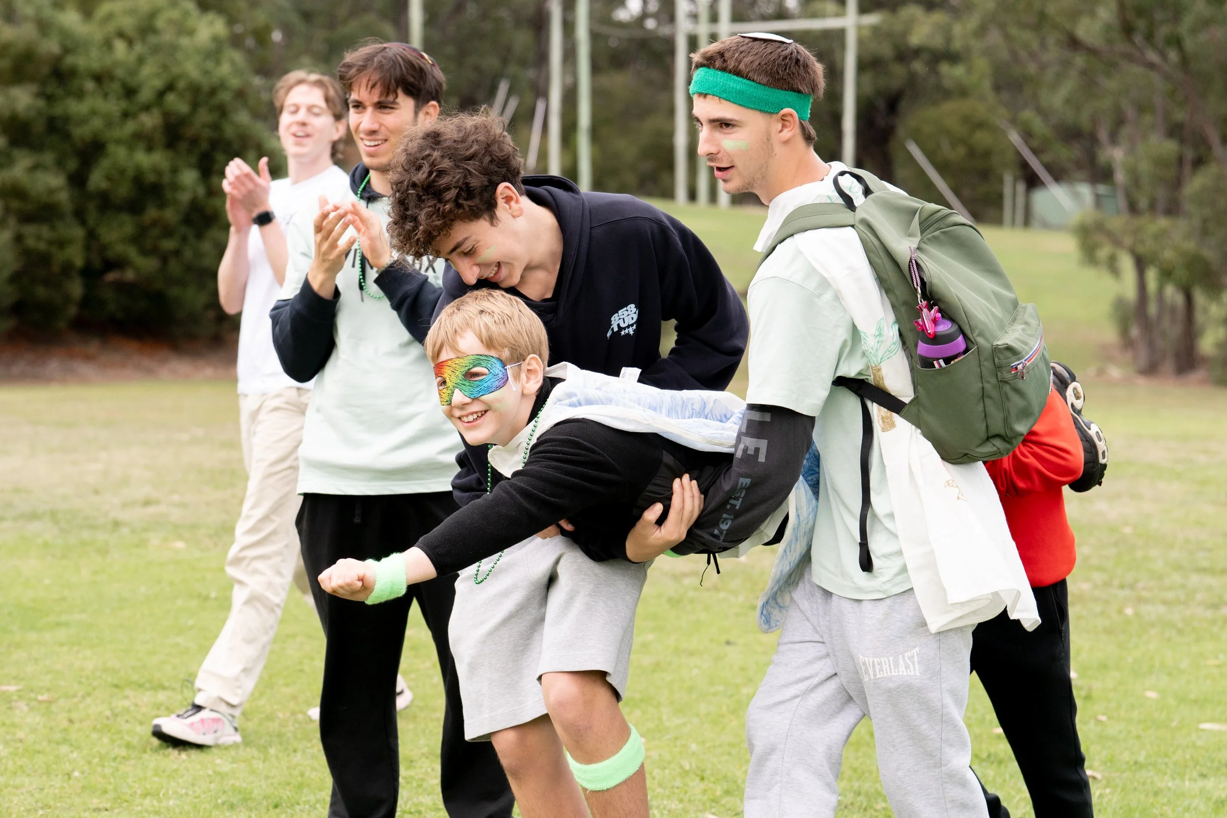 Group of young people playing outdoors, one person is being lifted and is wearing a colorful mask and a black hoodie.