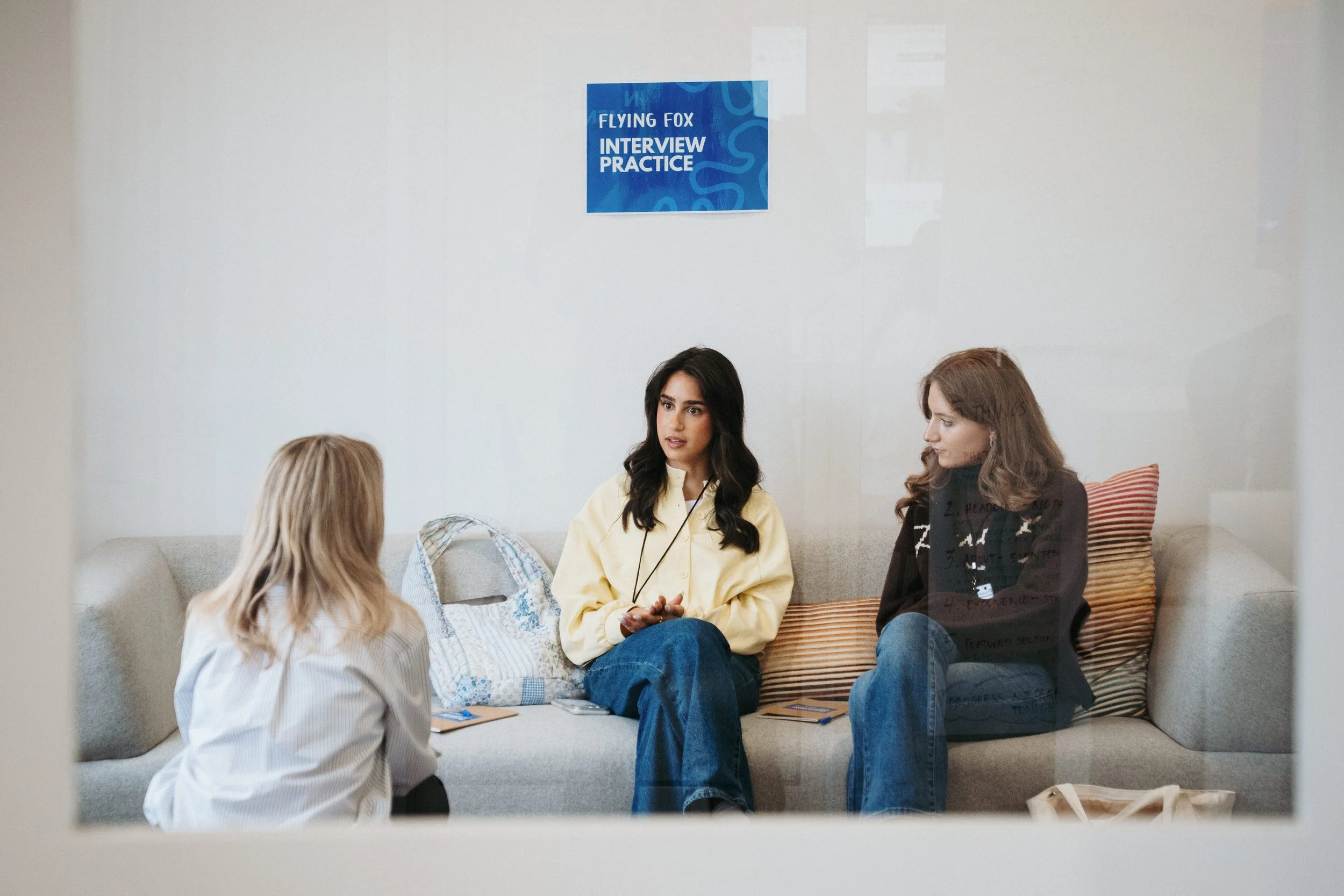 Three women having a conversation during a job interview in an office, with a sign on the wall that reads 'Flying Fox Interview Practice'.