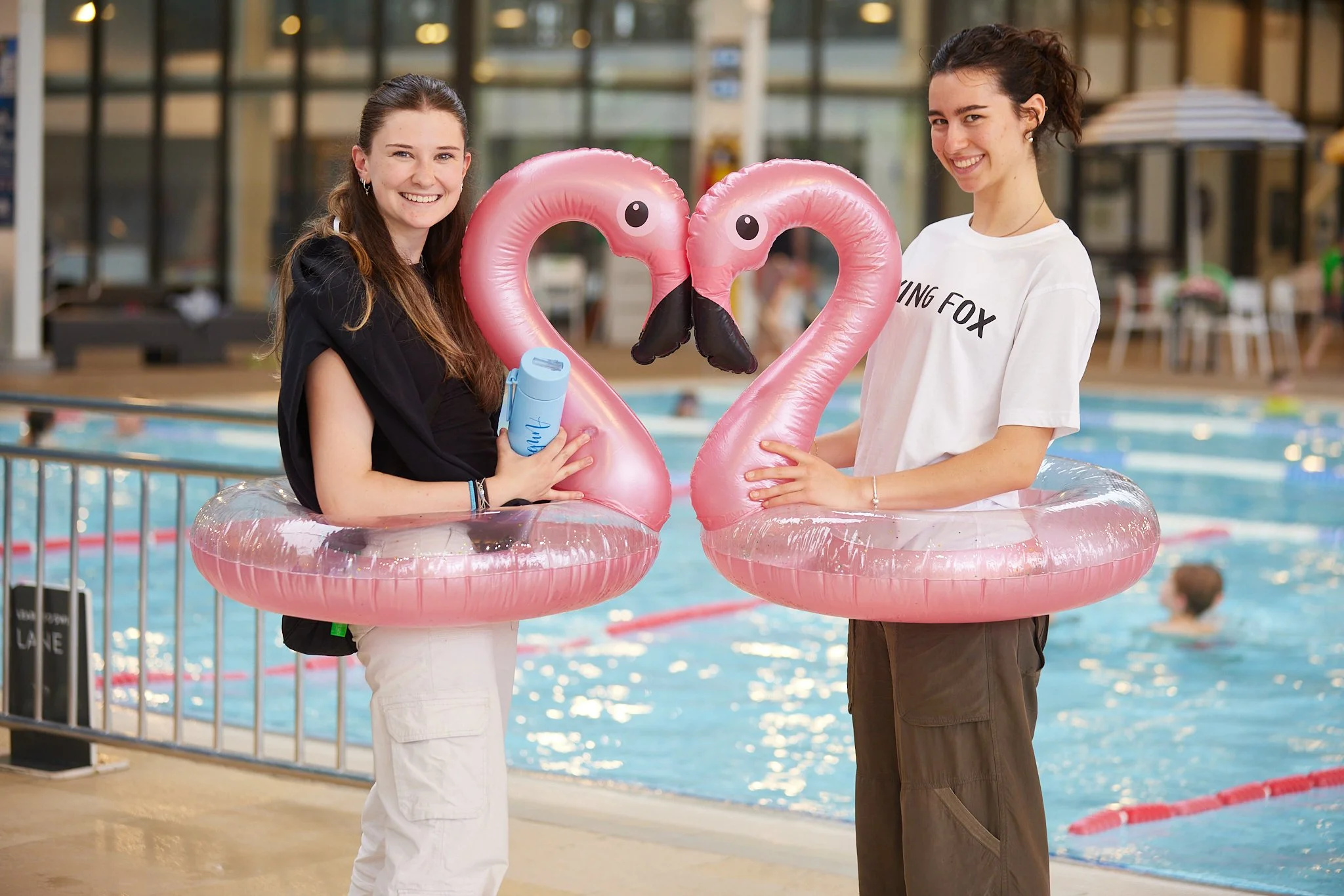 Two young women standing in an indoor swimming pool area, holding pink flamingo-shaped pool floats with a black beak and eyes, smiling at the camera.