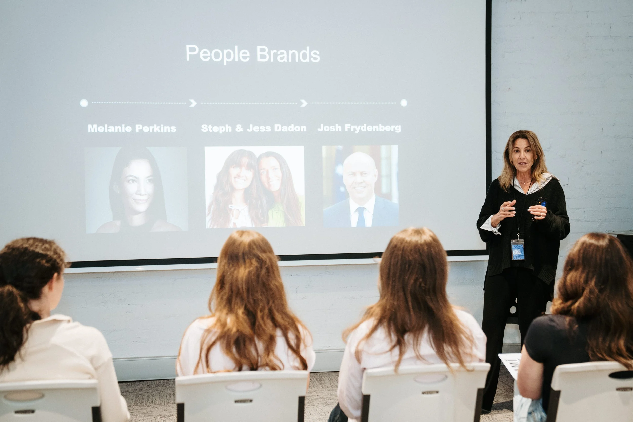 A woman giving a presentation in front of a slide that reads 'People Brands' showing photos and names of Melanie Perkins, Steph & Jess Dadon, and Josh Frydenberg, with four women seated facing her in a conference room.