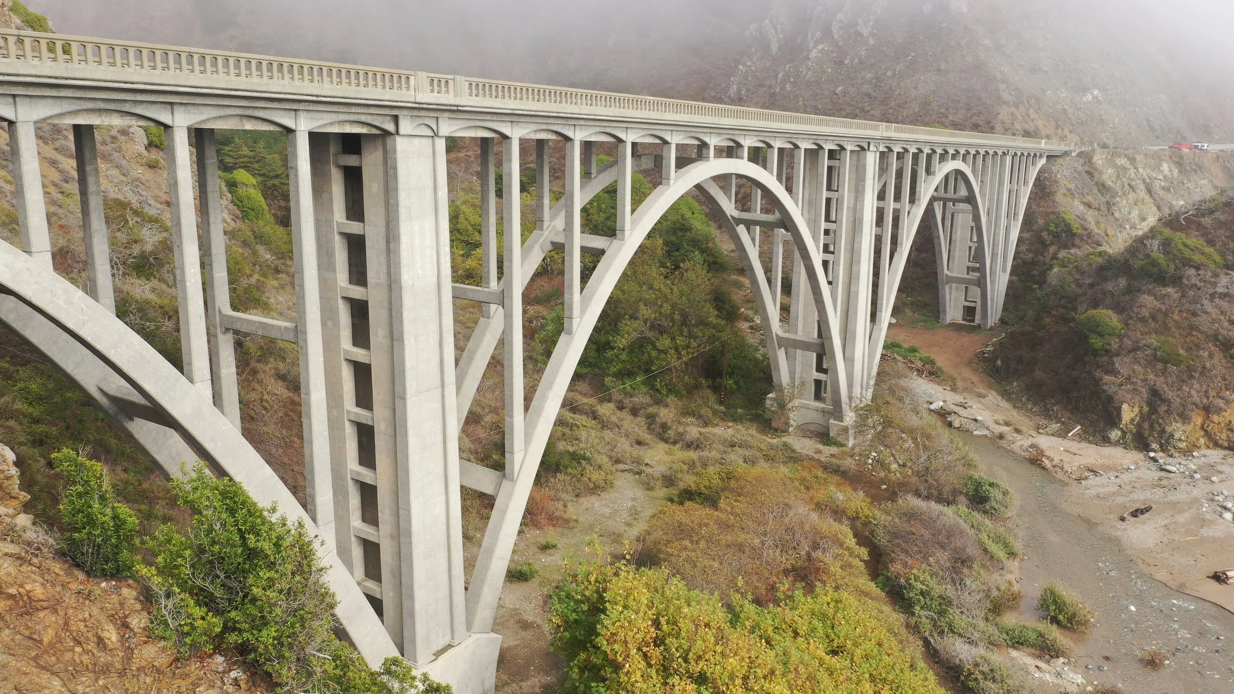 A large concrete arch bridge with multiple support columns crossing a dry riverbed in a mountainous area with fog.
