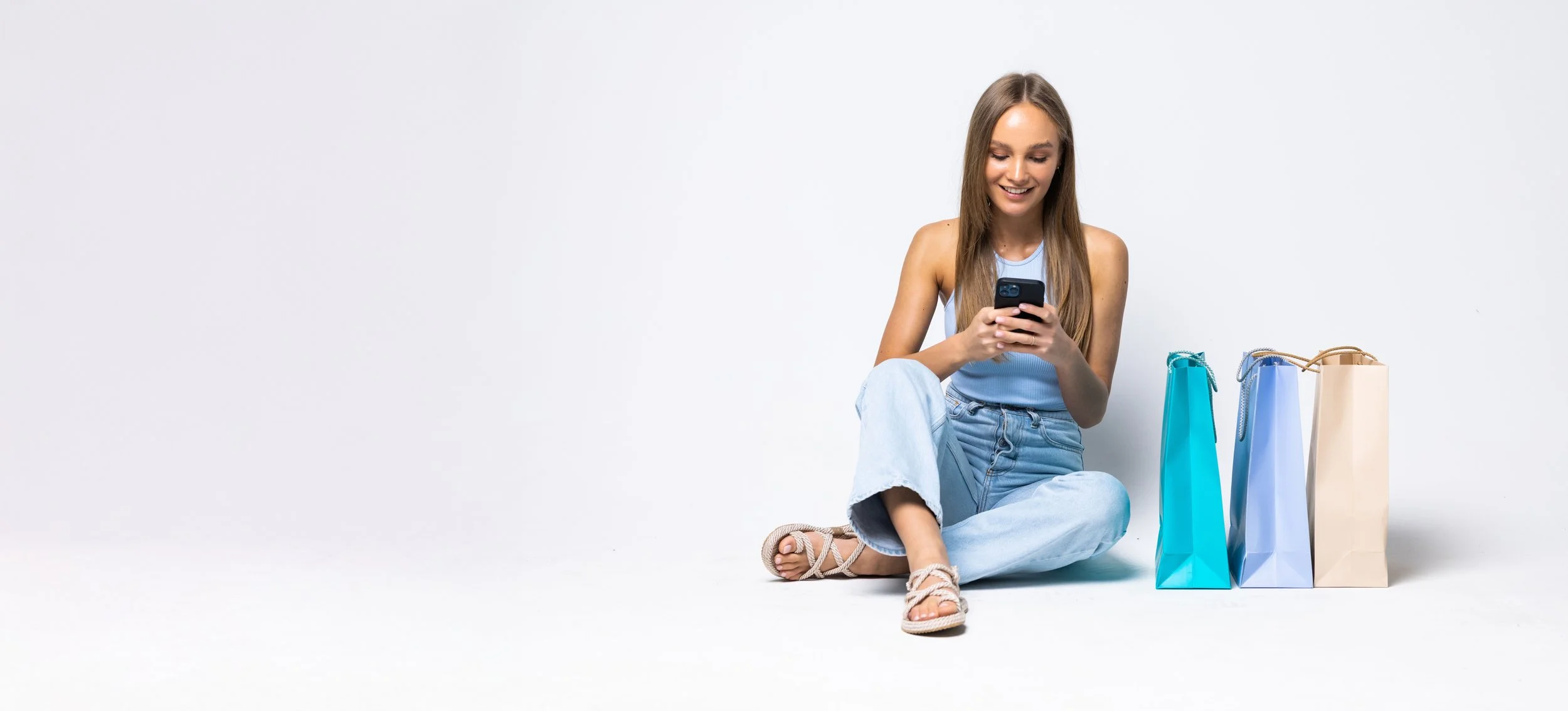 A young woman sitting on the floor, smiling while looking at her phone, with four shopping bags in different shades of blue and beige beside her, against a plain white background.