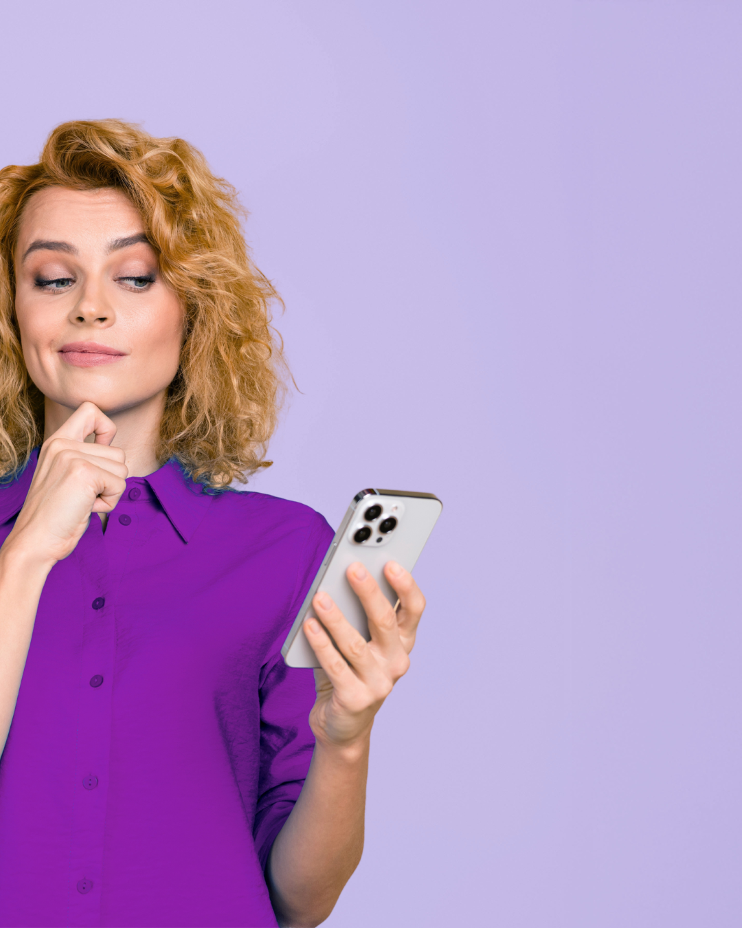A woman with curly red hair wearing a purple shirt looking at her smartphone with a neutral expression against a plain purple background.