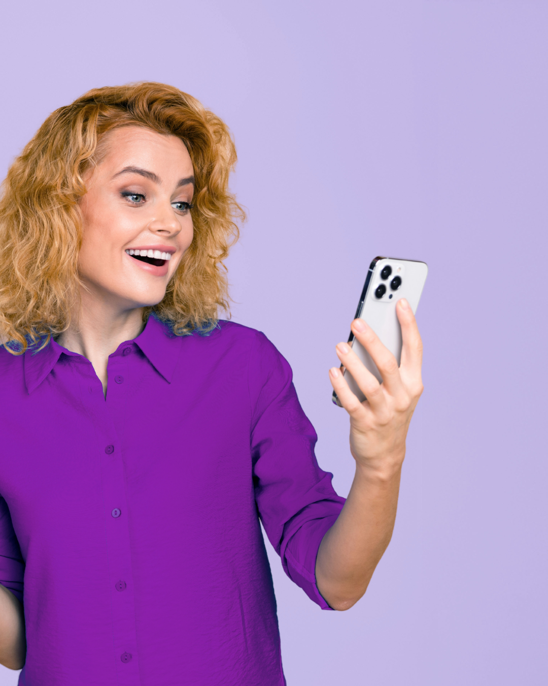 A woman with curly red hair wearing a purple shirt, smiling and looking at her smartphone against a purple background.