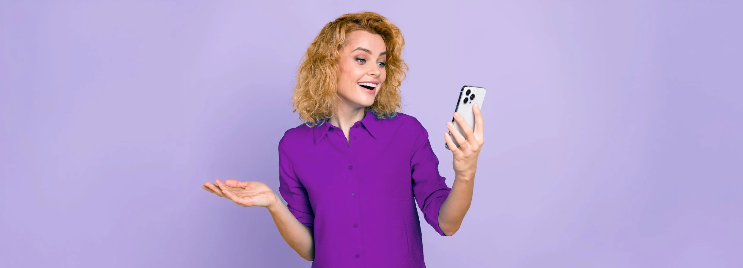 A woman with curly red hair wearing a purple shirt is looking at her phone with an excited expression, standing against a light purple background.