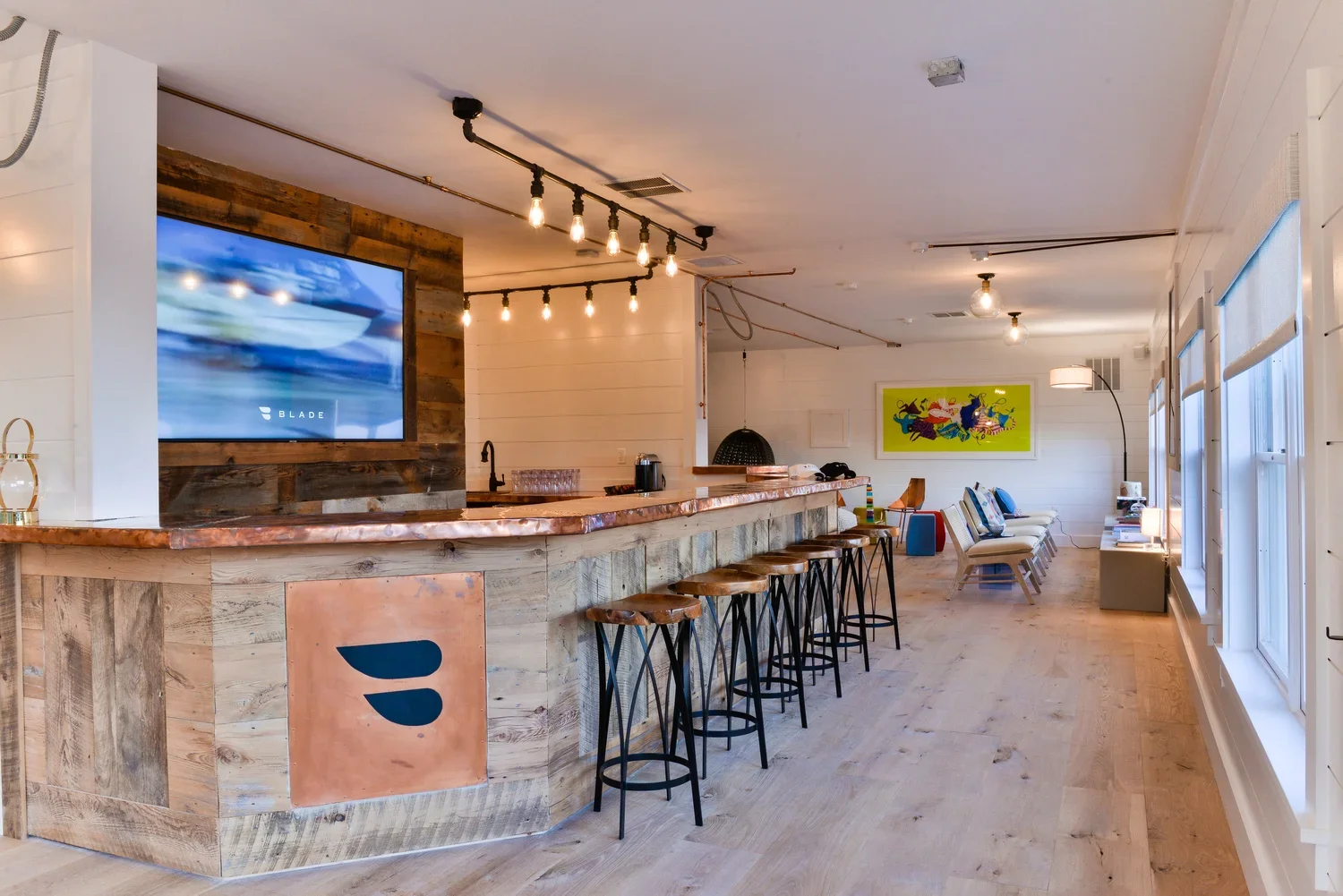 Modern lounge area with a bar counter, wooden stools, wall-mounted TV, and seating area with chairs and a colorful painting on white walls.