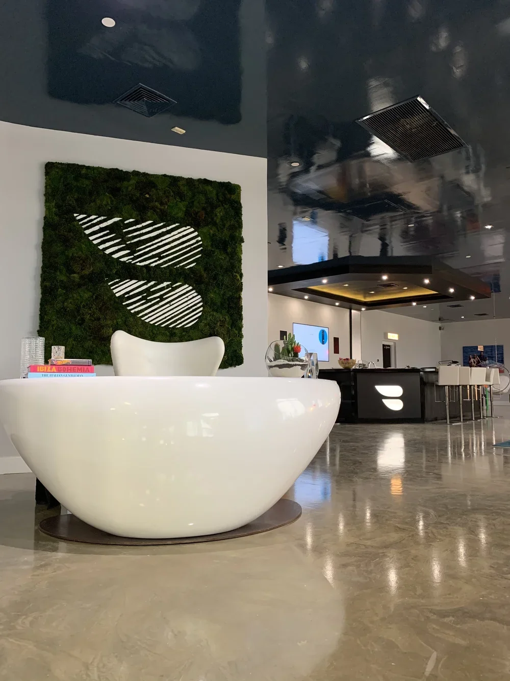 Modern hotel lobby with a white curved reception desk, green moss wall art, and a glossy black ceiling with recessed lighting.