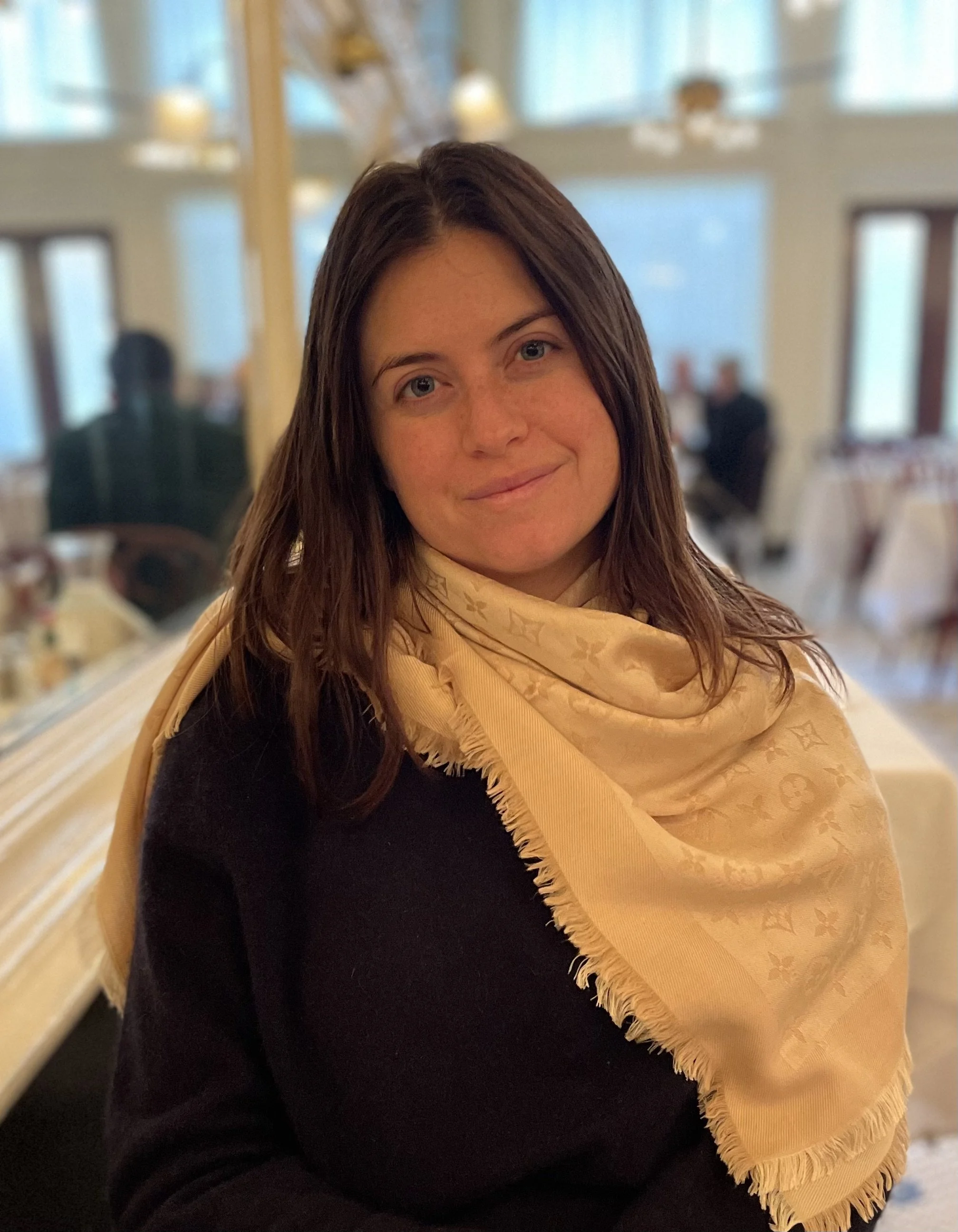 A woman with shoulder-length brown hair, wearing a black top and a light beige scarf, sitting in a restaurant with blurred background of tables and people.
