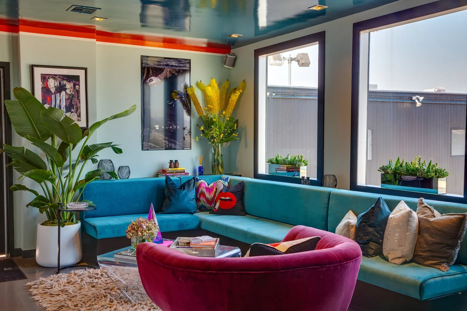 Living room with a large teal sectional sofa, a pink velvet armchair, a glass coffee table, and various decorative pillows. A large potted plant, framed artwork, and windows with planter boxes filled with succulents are visible.