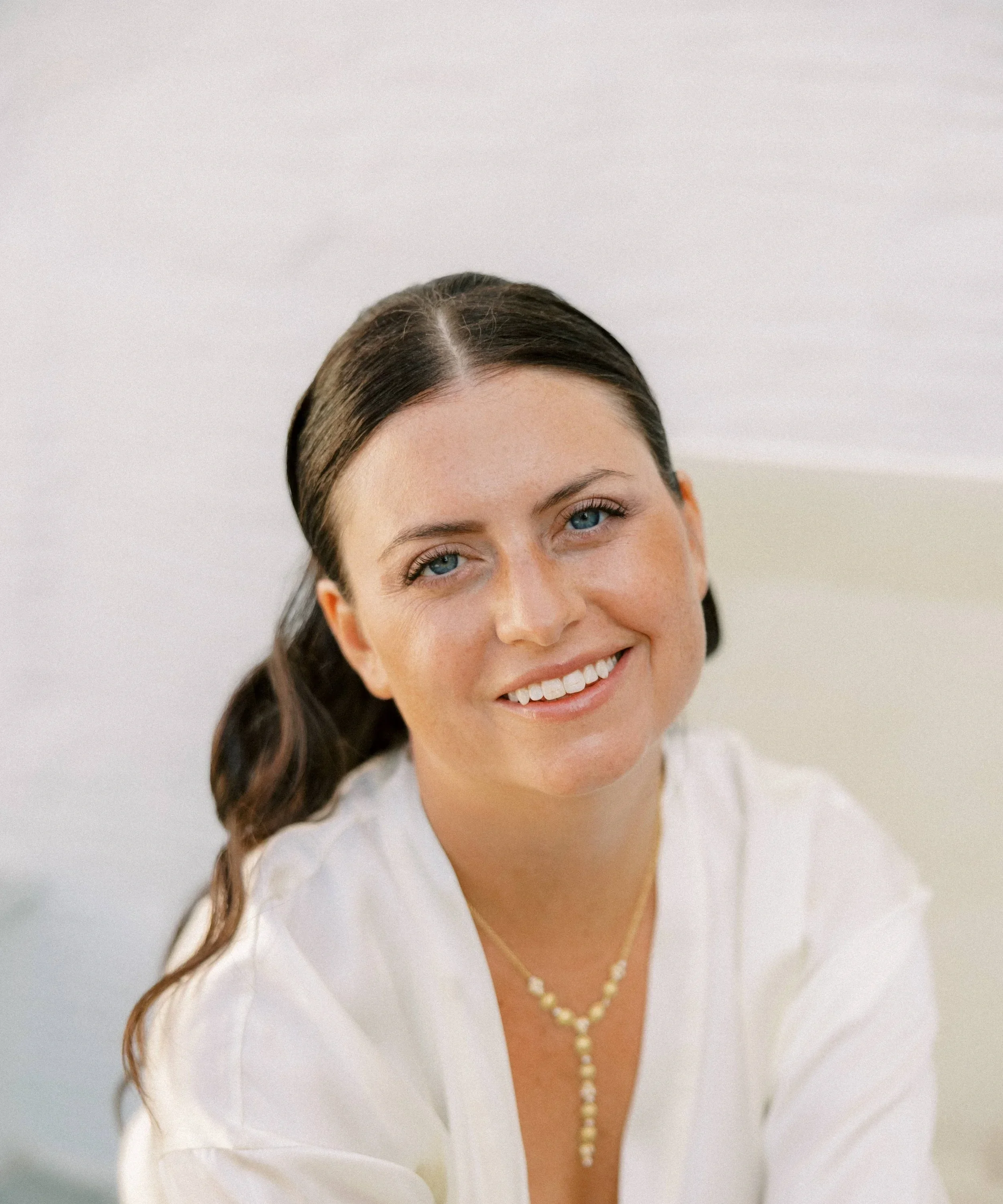 Close-up portrait of a smiling woman with long dark hair, blue eyes, and light skin, wearing a white top and a pearl necklace.
