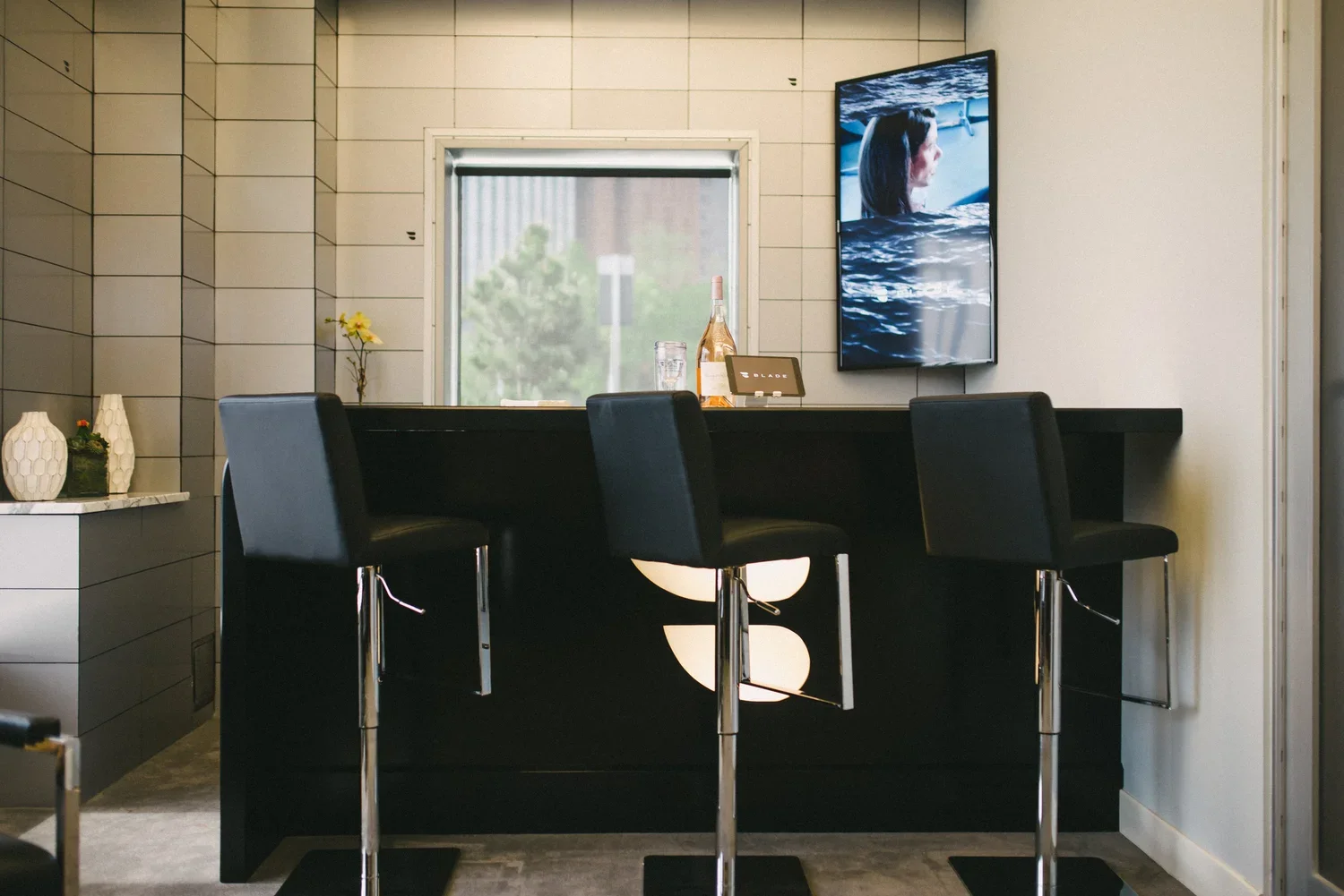 A modern indoor bar with three black bar stools facing a black bar counter, a window behind with greenery outside, a flat-screen TV mounted on the wall showing a woman with long dark hair, and decorative vases on a counter to the left.