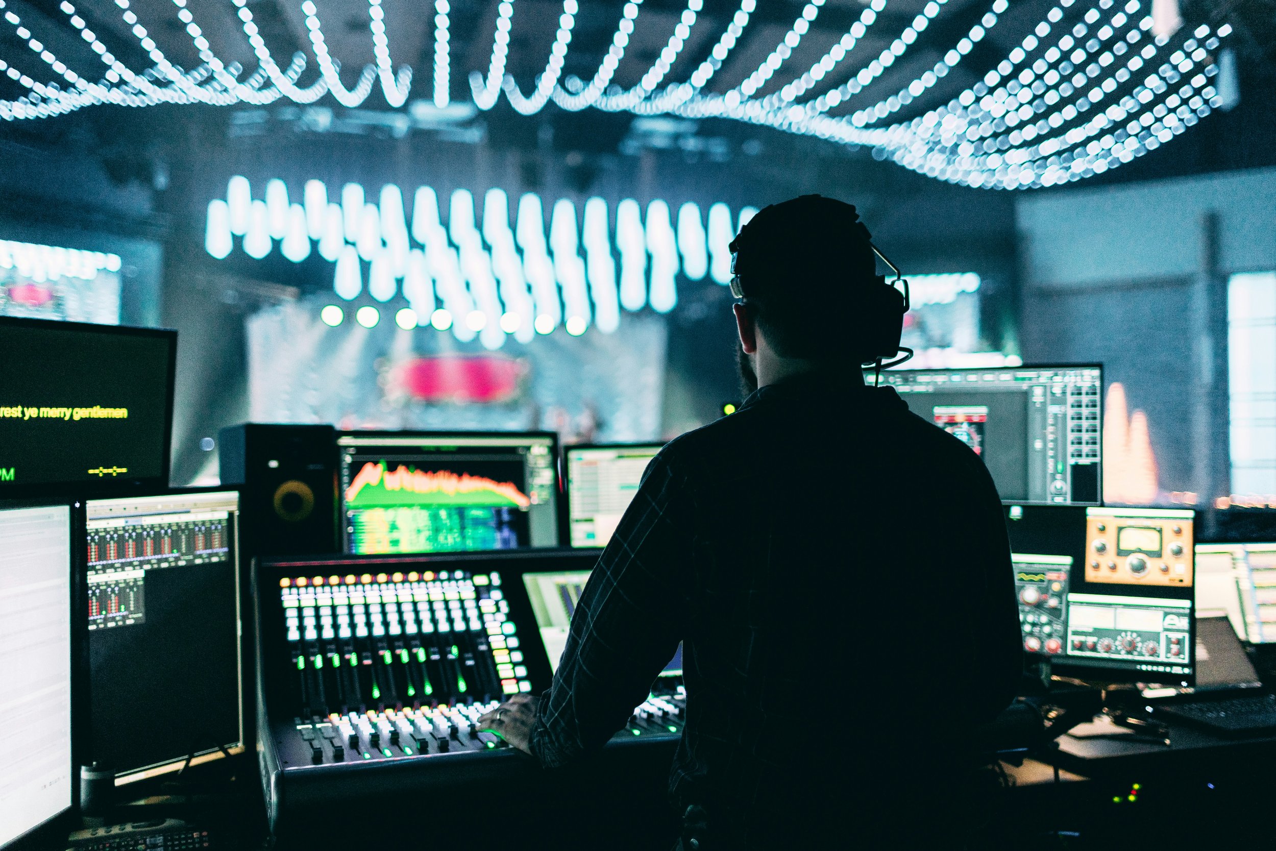 Silhouette of a DJ working with multiple computer screens and audio equipment in a dark room with bright, colorful lights and a blurred stage or performance area in the background.