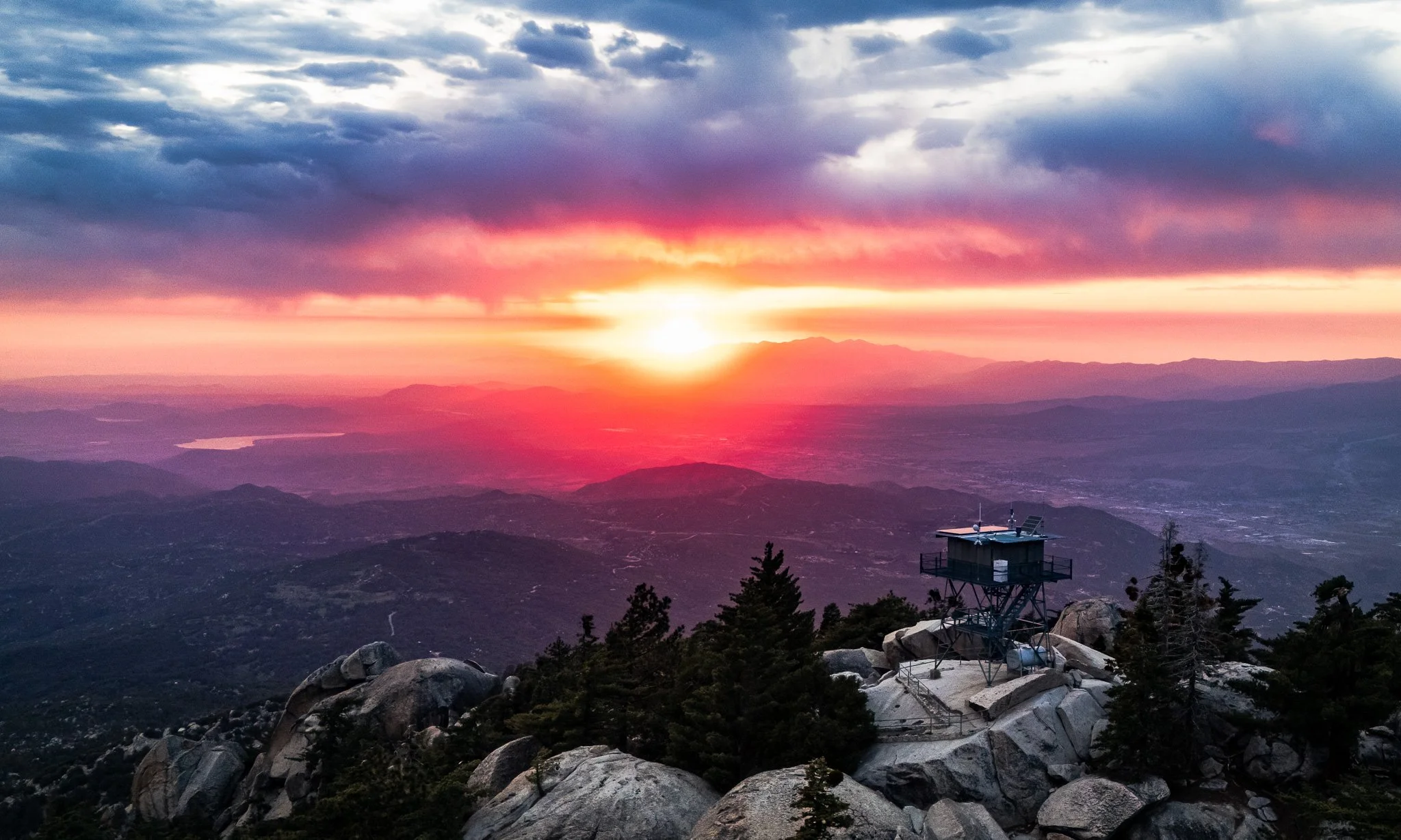 Sunset over mountains with colorful clouds, including pink and purple hues, viewed from a rocky hilltop with a small observation station surrounded by trees.