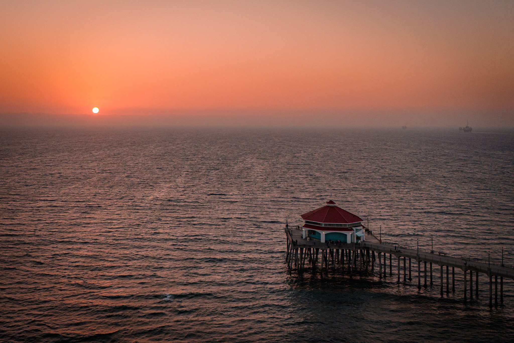 Sun setting over the ocean with a pier and small building at the end, pink and orange sky, calm water, ships visible in the distance.