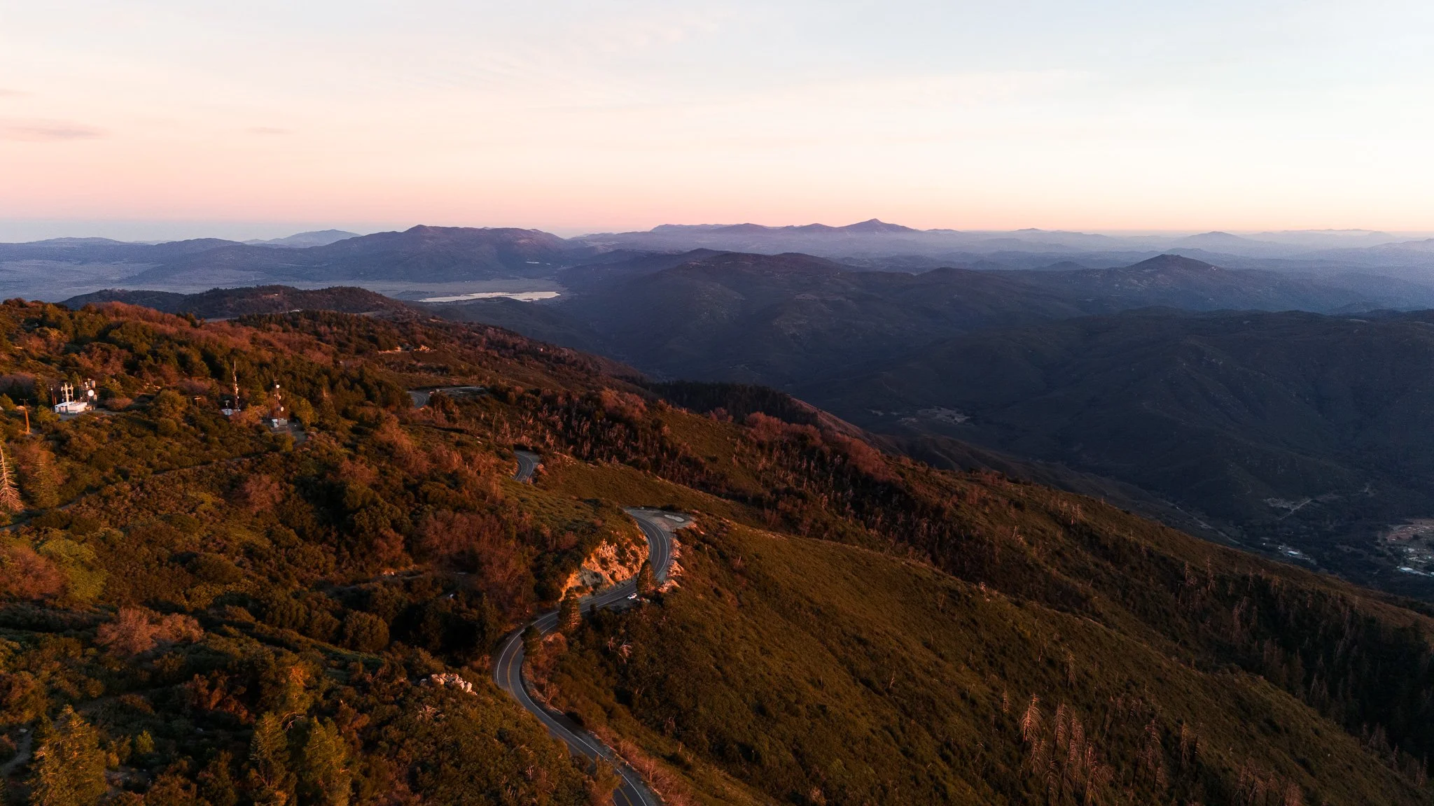 A scenic view of a mountain landscape at sunset with a winding road through green and brown hills, distant mountain ranges, and a partly cloudy sky.
