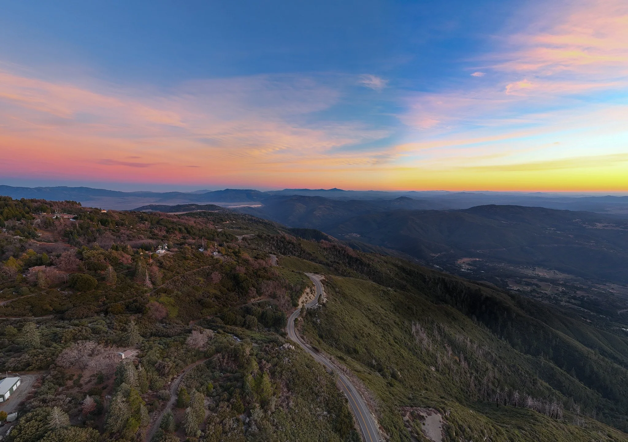 Aerial view of a winding mountain road through green hills and valleys during sunset with colorful sky