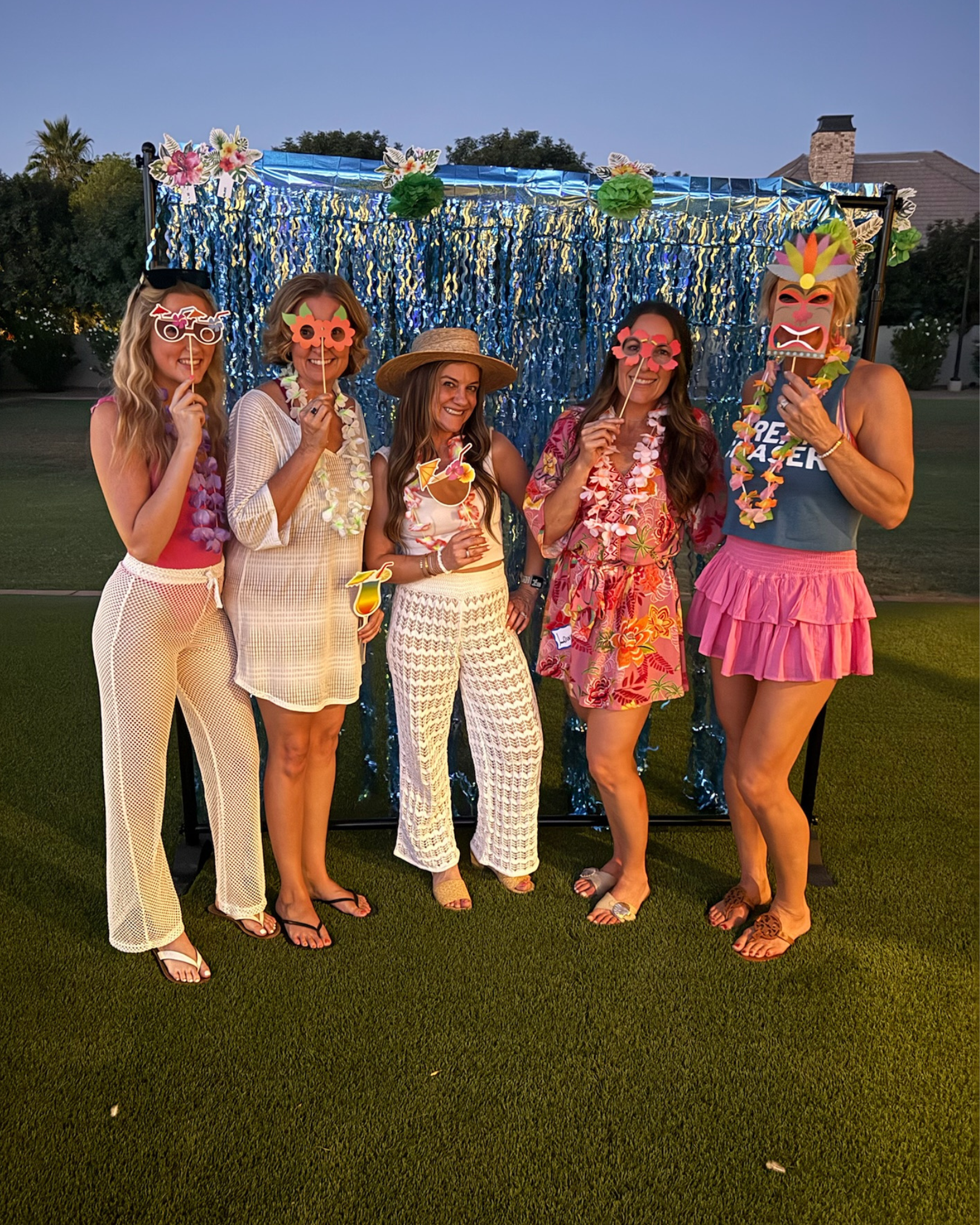 Five women in colorful summer outfits posing outdoors at a tropical-themed party backdrop, holding fun tiki mask props.