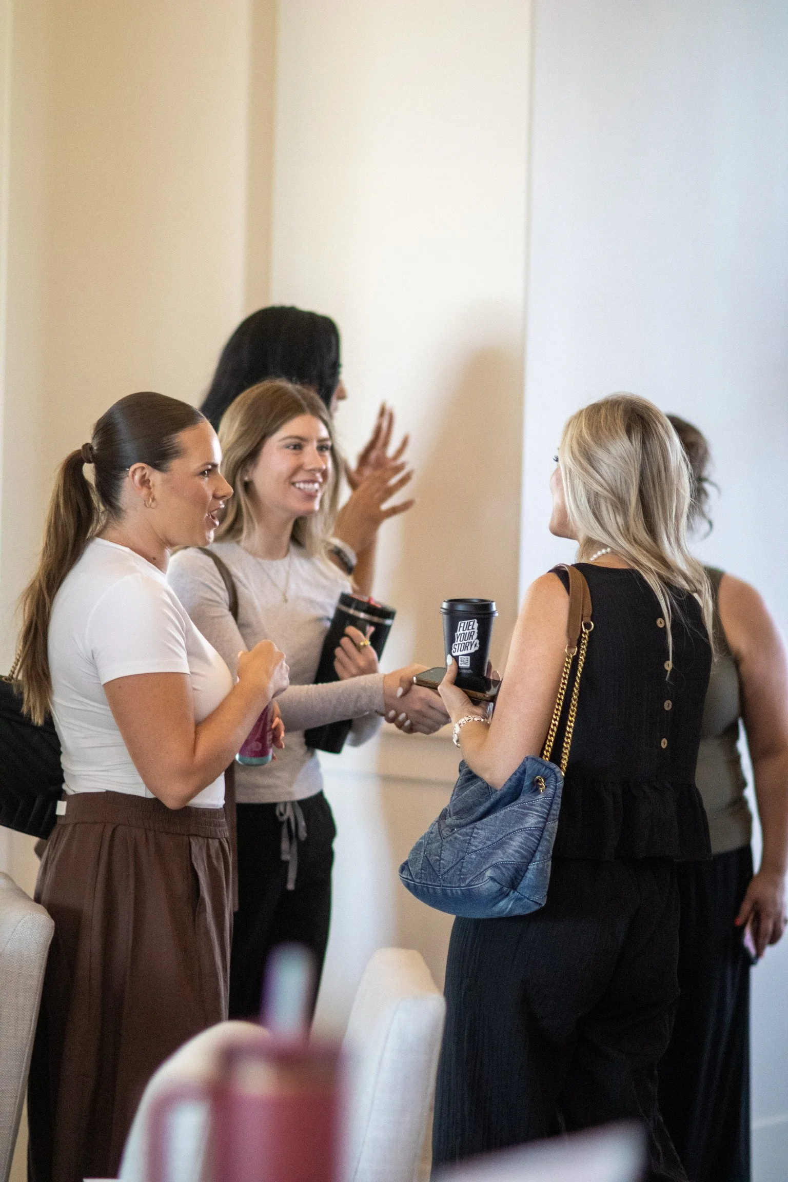 A group of women engaging in conversation indoors, some holding coffee cups, with a plain wall background.