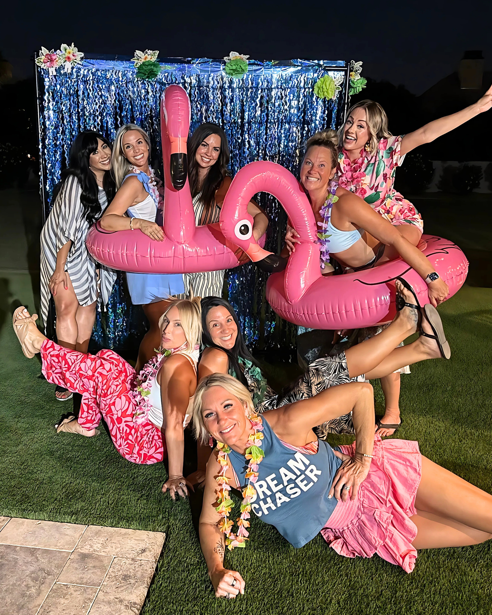 Group of women enjoying a summer party with inflatable pink flamingo pool floats, wearing colorful clothes and leis, posing in front of a blue sequin backdrop outdoors at night.