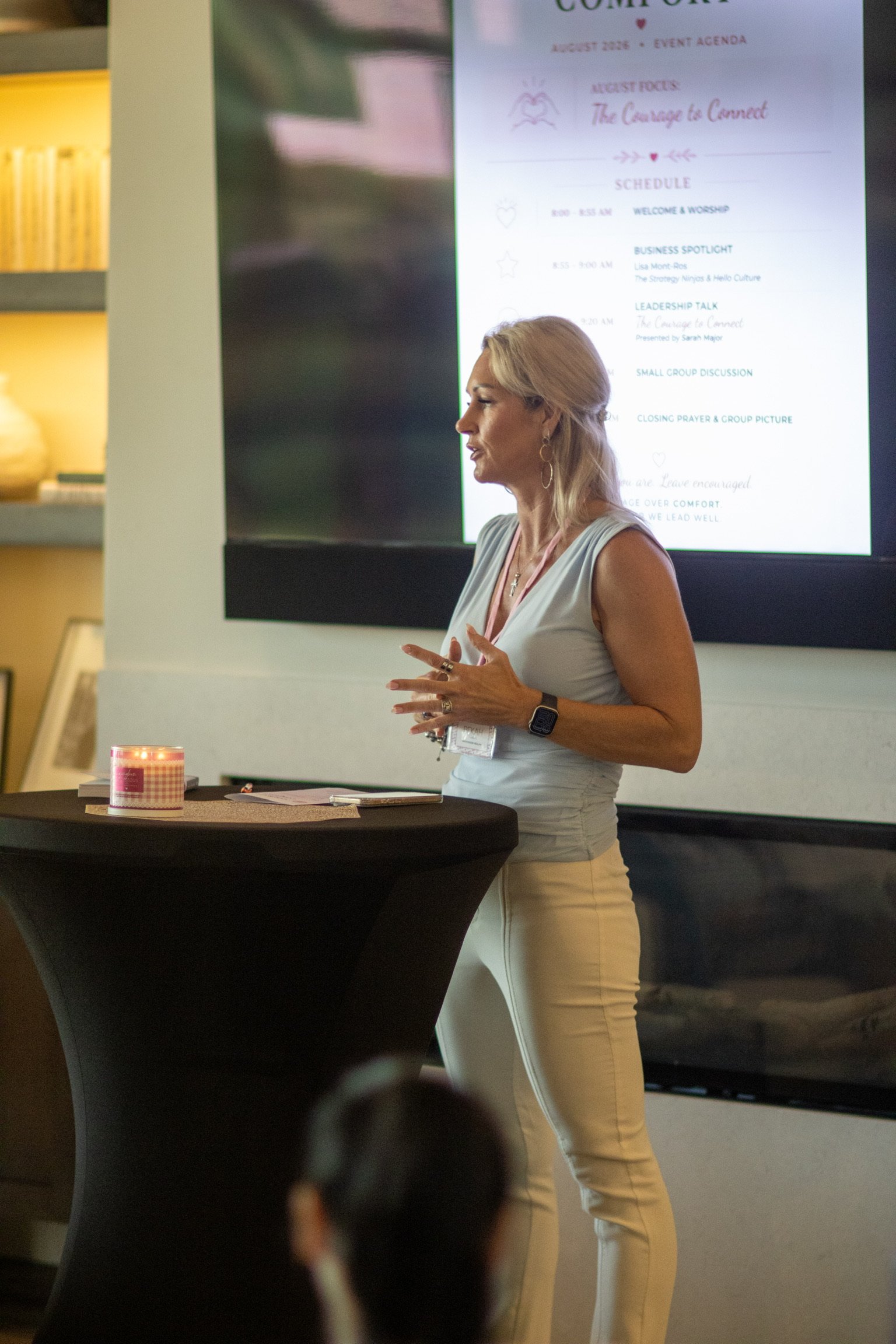 A woman with blonde hair speaking at a presentation, standing beside a large screen displaying an event schedule, while a candle is on a table in the foreground.