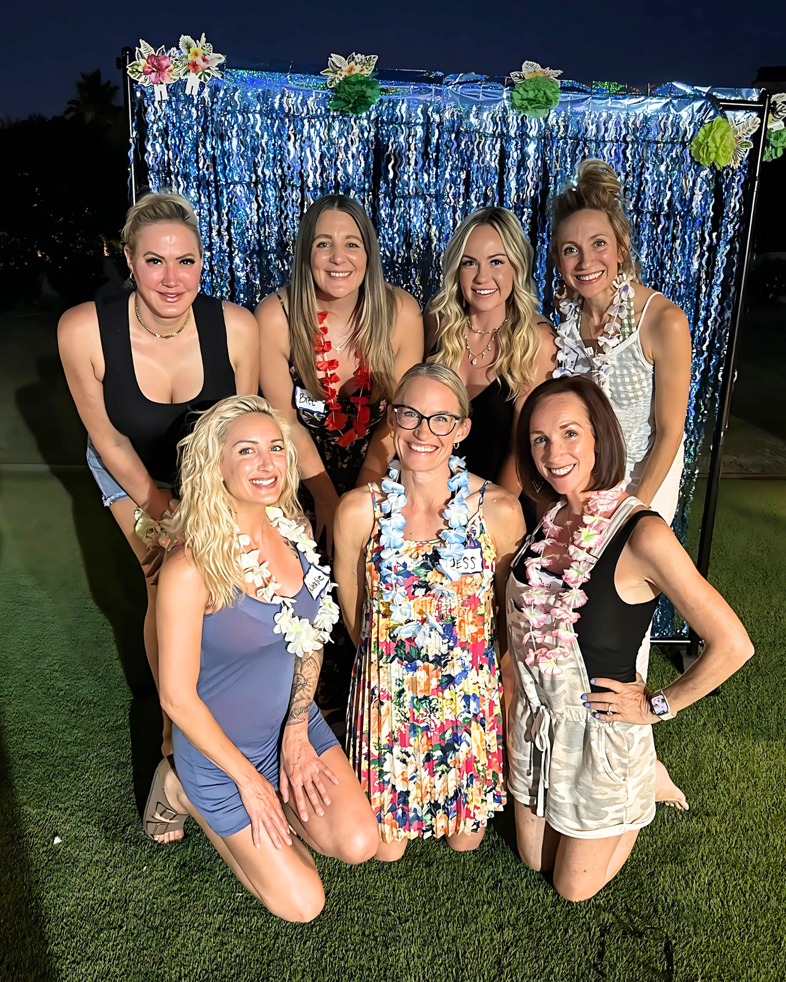 Group of nine women wearing leis, smiling at a nighttime outdoor party with a decorated backdrop of blue shimmering curtains and paper flowers.