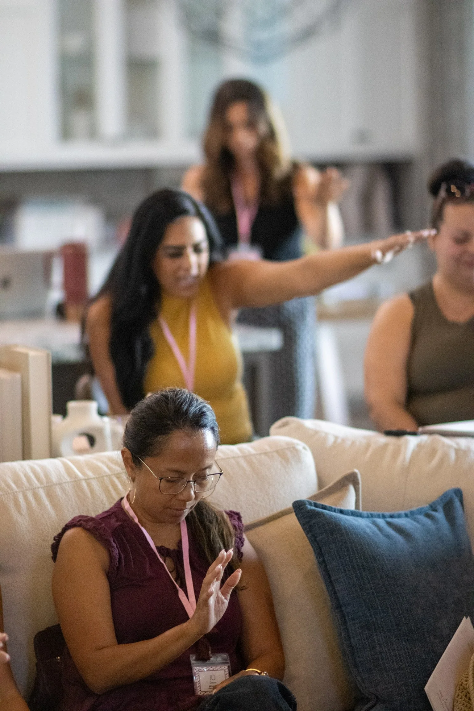 A group of women in a casual setting, some sitting on a beige sofa and others standing behind, engaging with their phones and each other in a well-lit room with shelves in the background.