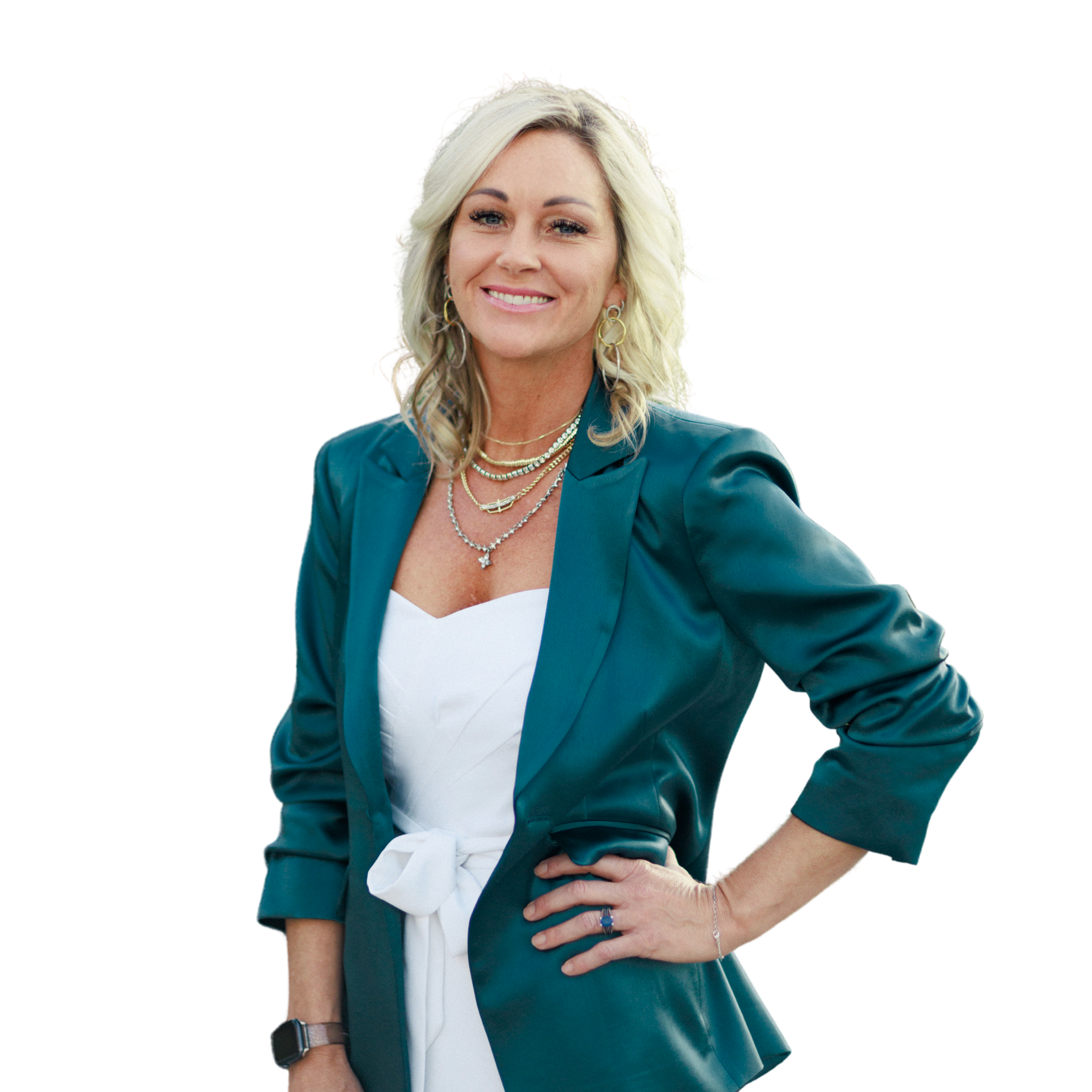 A blonde woman with shoulder-length hair, smiling, wearing a teal blazer over a white top tied at the waist, layered necklaces, earrings, and a watch, standing with one hand on her hip.