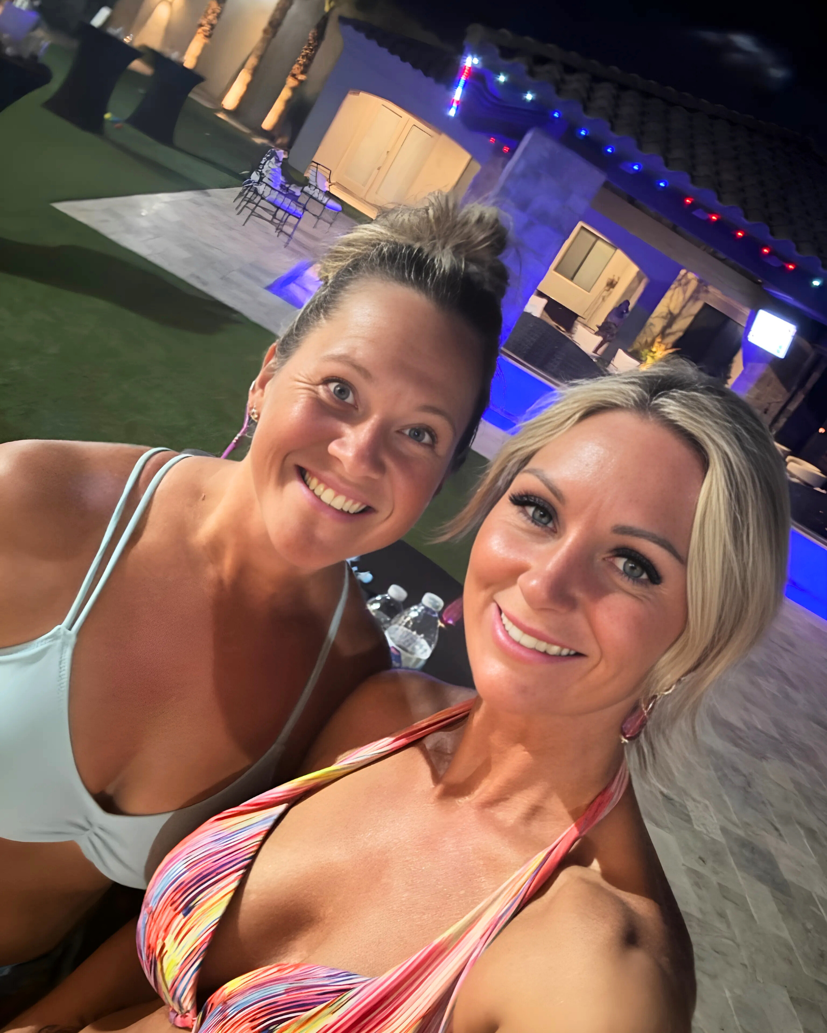 Two women taking a selfie at night outdoors, with a house decorated with string lights in the background.