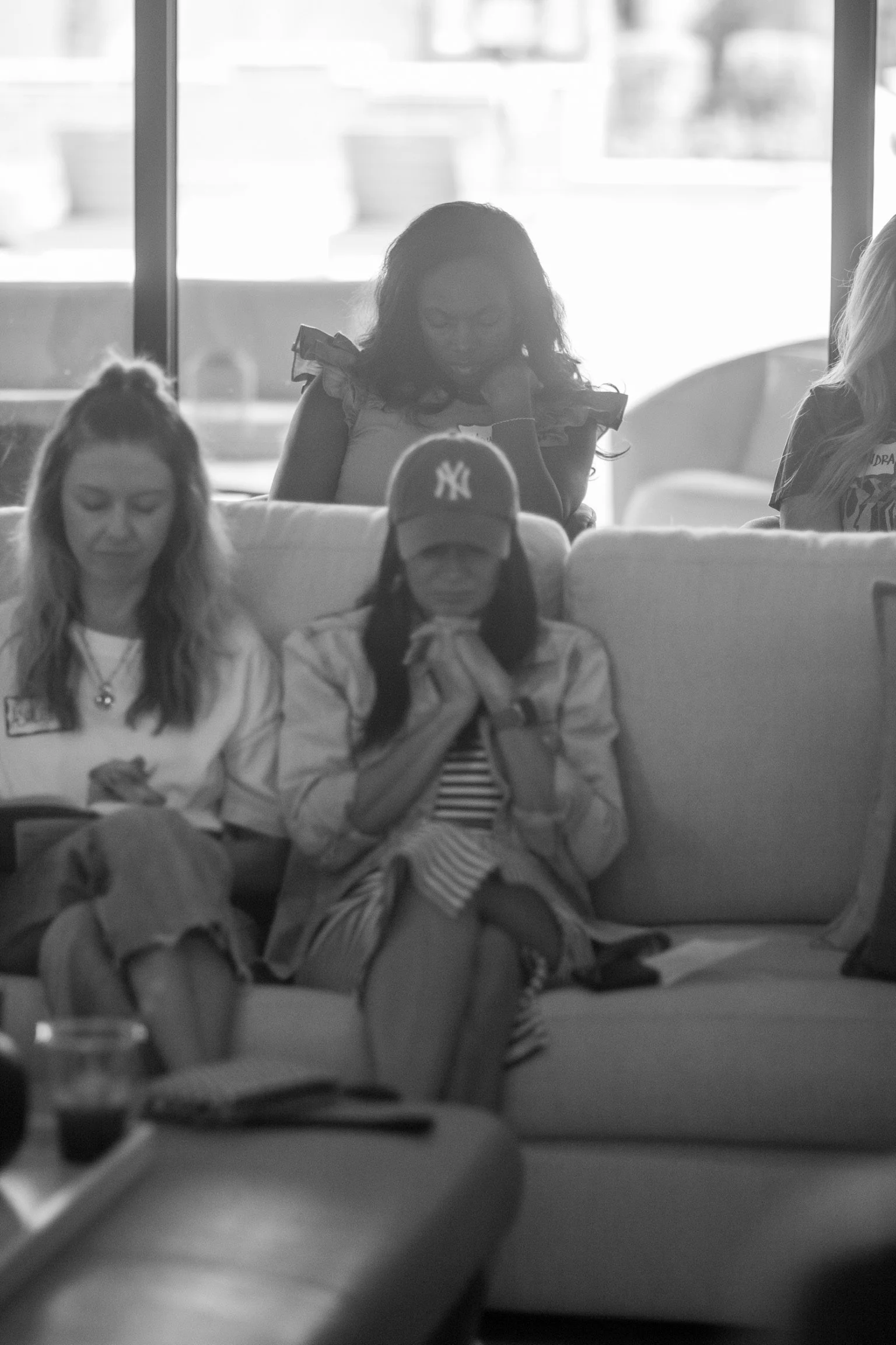 Black and white photo of four women sitting on a sofa in a living room. Two women are in the foreground, one with light hair and the other with dark hair wearing a baseball cap. The two women in the background are looking down, one with glasses and o