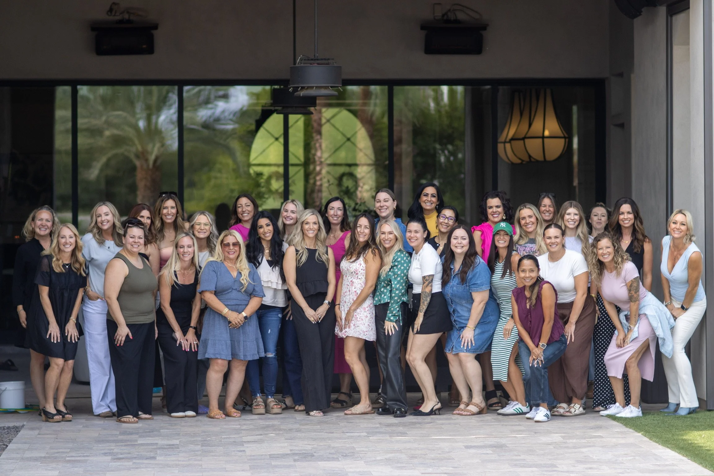A large group of women posing together outdoors in front of a building with large glass windows, some trees visible through the windows. The women are smiling and standing closely together, dressed in casual and semi-formal clothing.