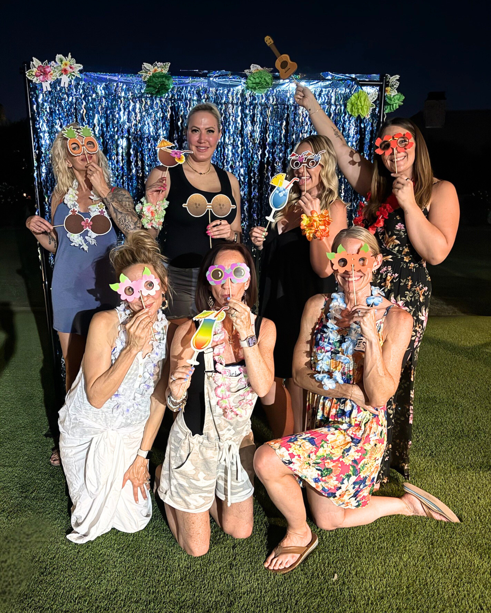 Group of women at a party wearing colorful sunglasses and leis, holding tiki drink props, with a blue sequin backdrop and Hawaiian decorations.