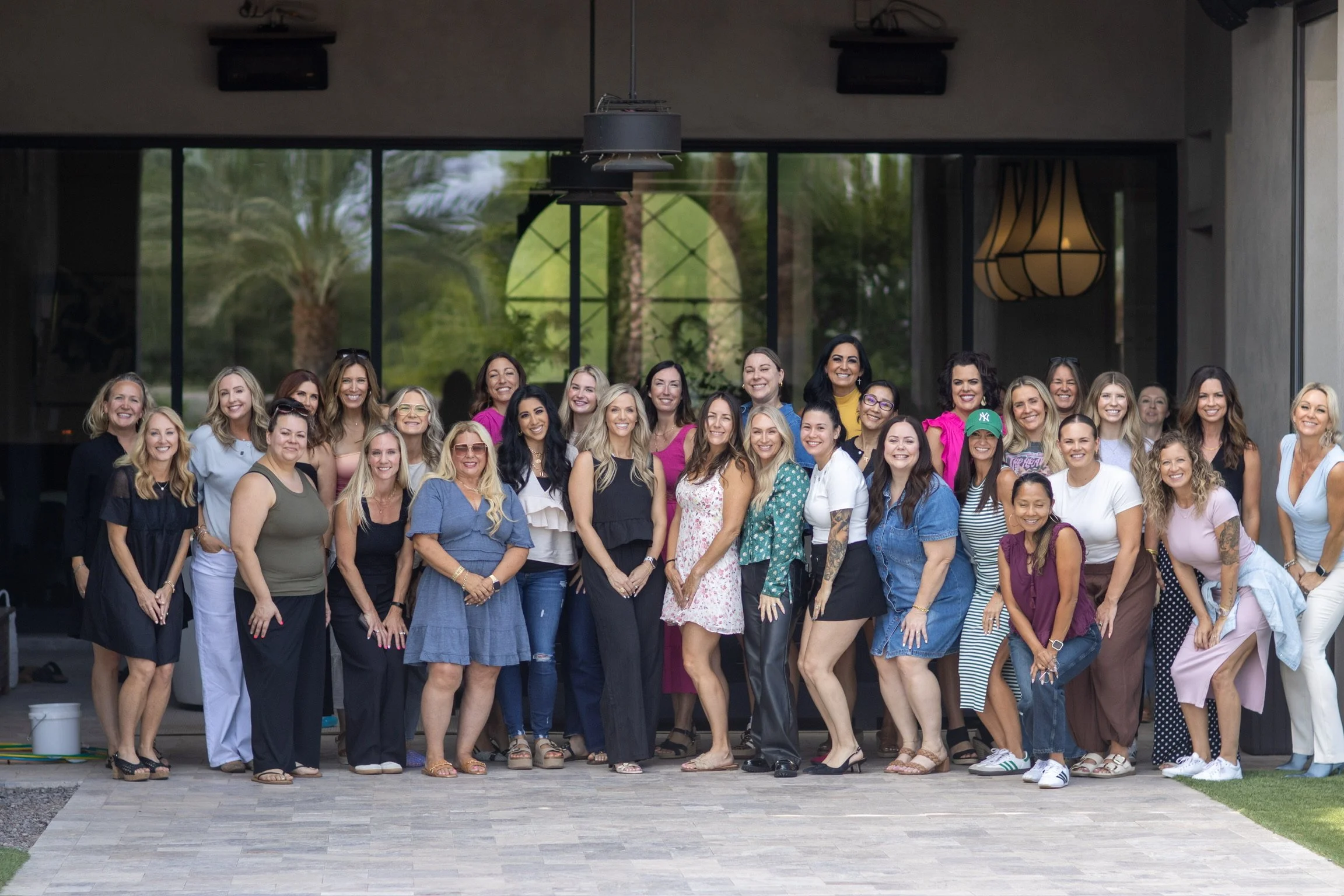 A large group of women standing together outdoors in front of a building with large windows and greenery. They are smiling and posing for a group photo.