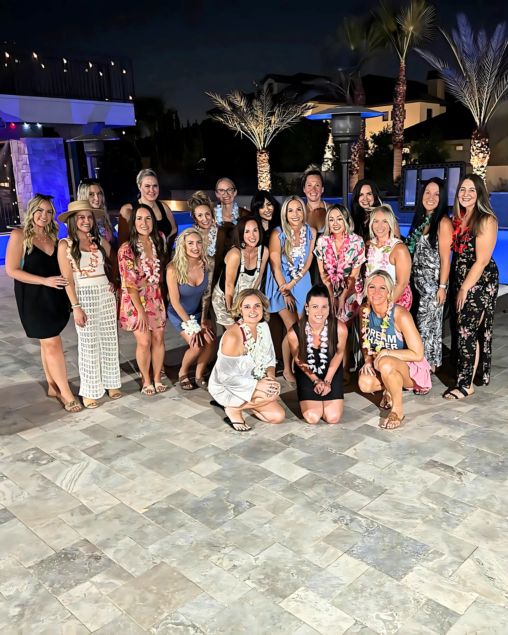 Group of women at a tropical-themed outdoor party at night, dressed in summer and Hawaiian shirts, some with leis, smiling in front of a swimming pool and palm trees.