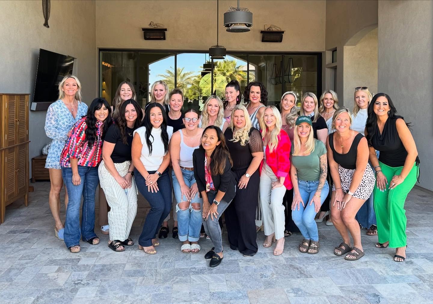 Group of women posing outdoors on a patio with a large glass window and palm trees in the background.