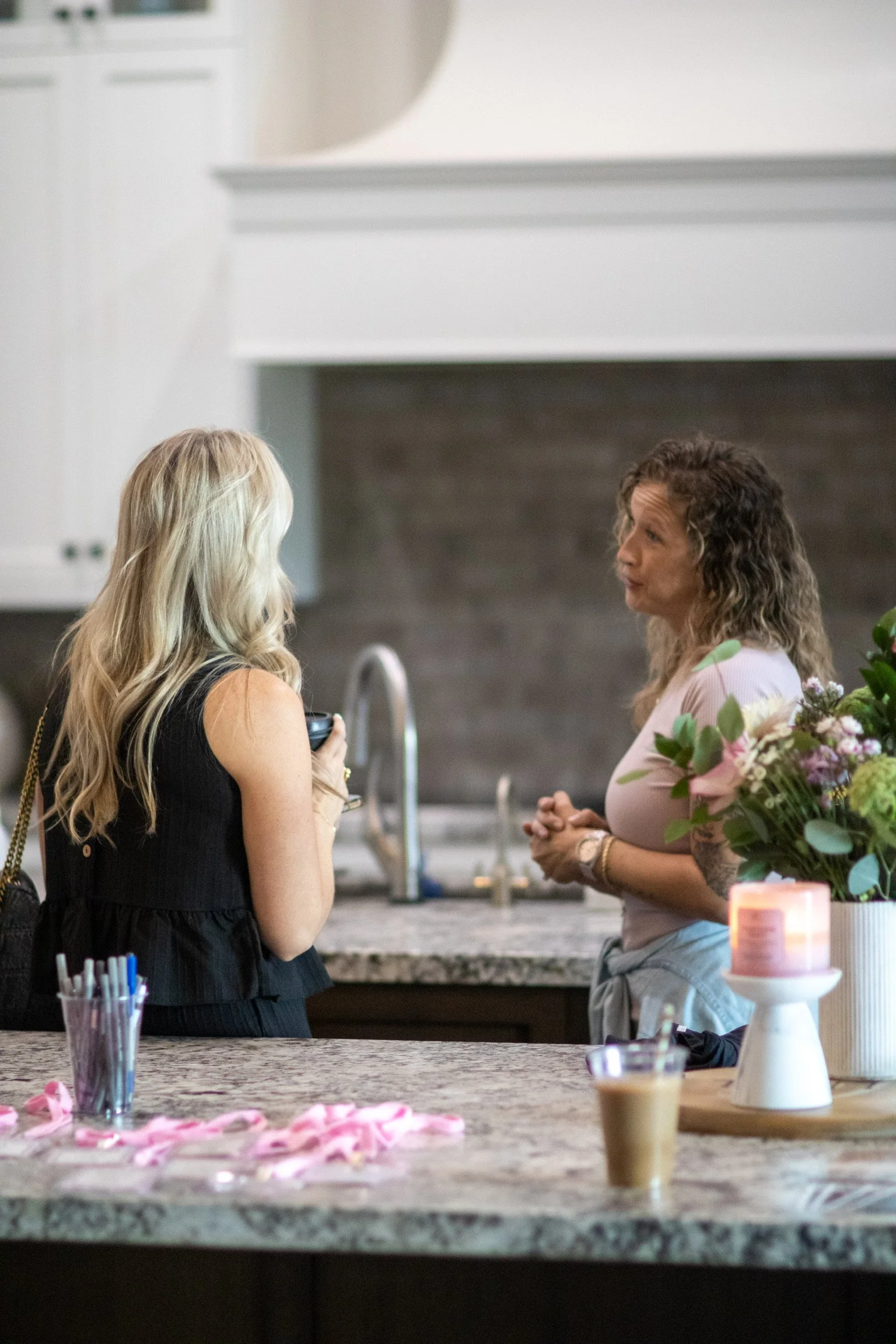 Two women are having a conversation in a kitchen near a counter, with pink decorations and flowers on the counter.